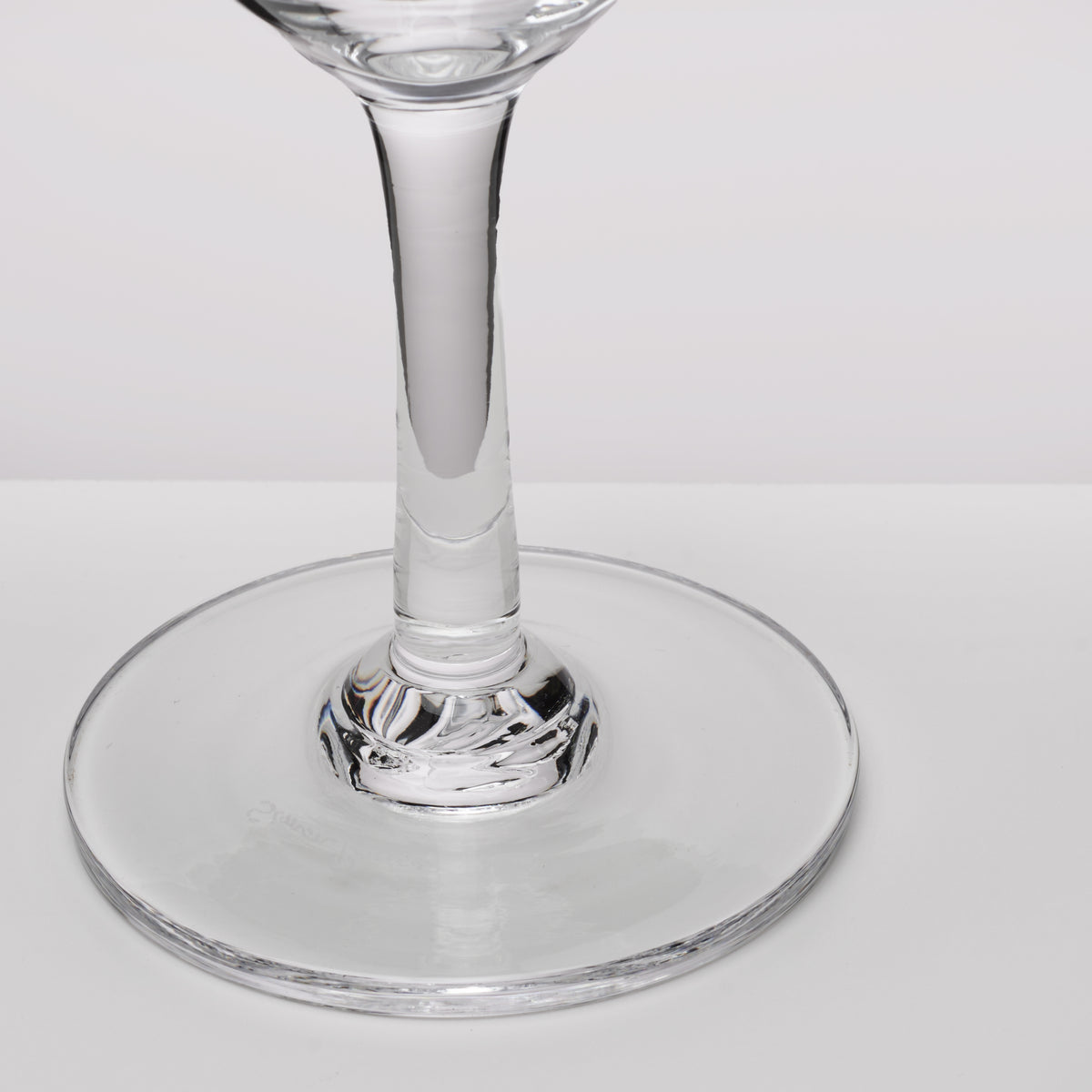 A close-up of a clear glass wine glass showing the stem and circular base against a light gray background.