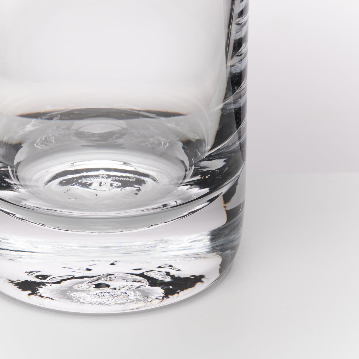 A close-up of a clear glass tumbler with a thick, rounded base and subtle etched or textured detailing on the side, shown against a clean white background.