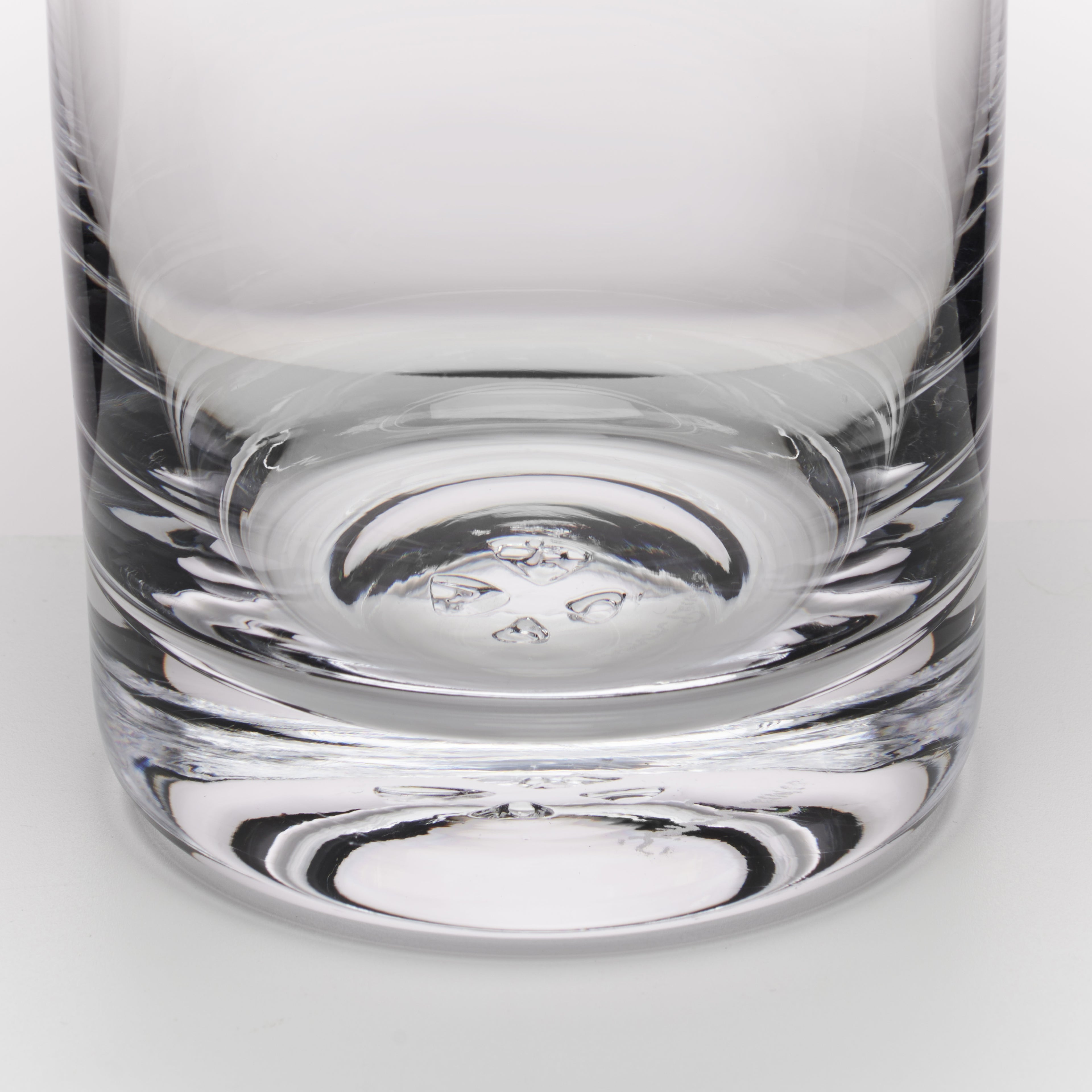 A close-up of a clear glass tumbler with a thick, textured base, showing reflections on a light gray background.