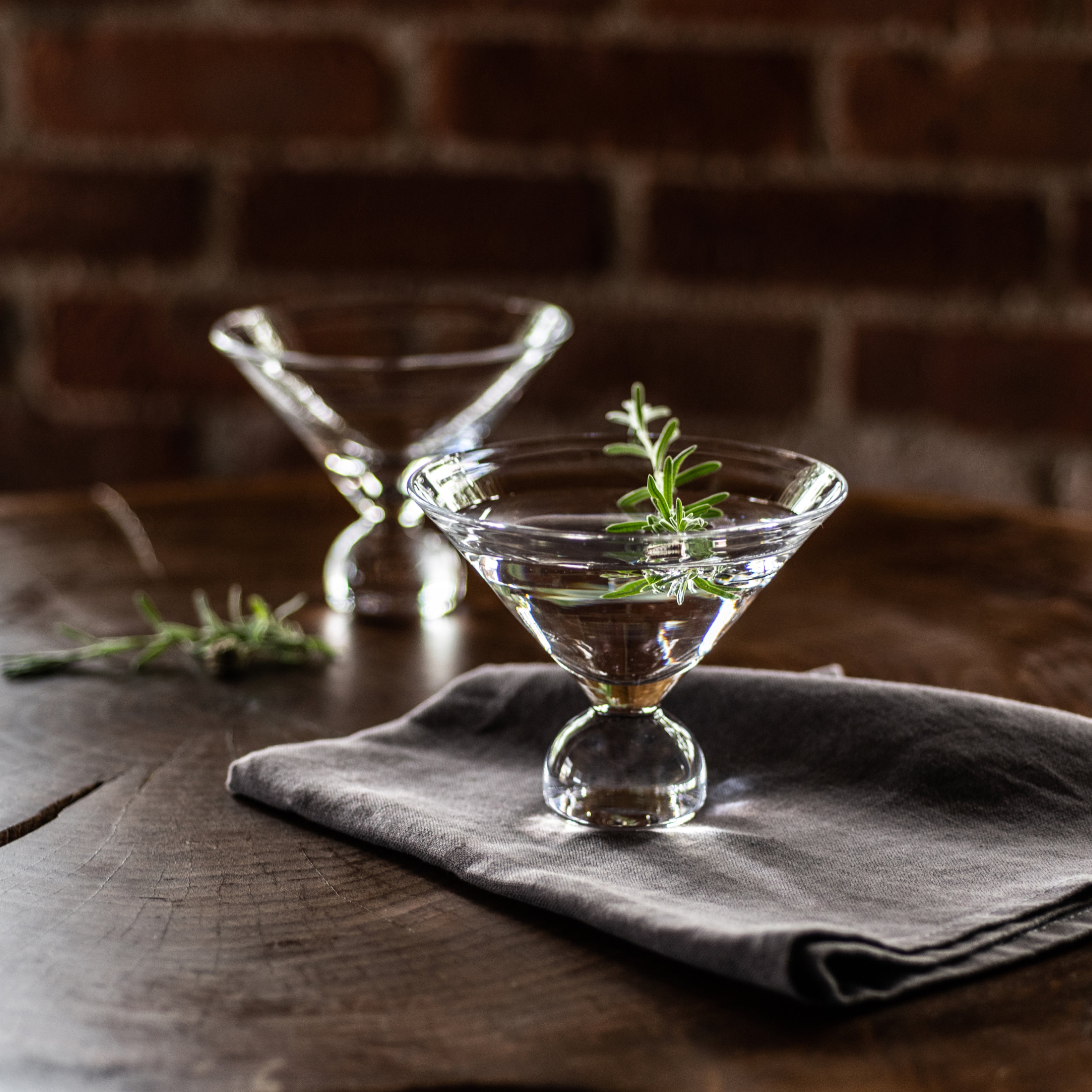 Two clear martini glasses with rosemary garnish on a gray napkin against a brick background.
