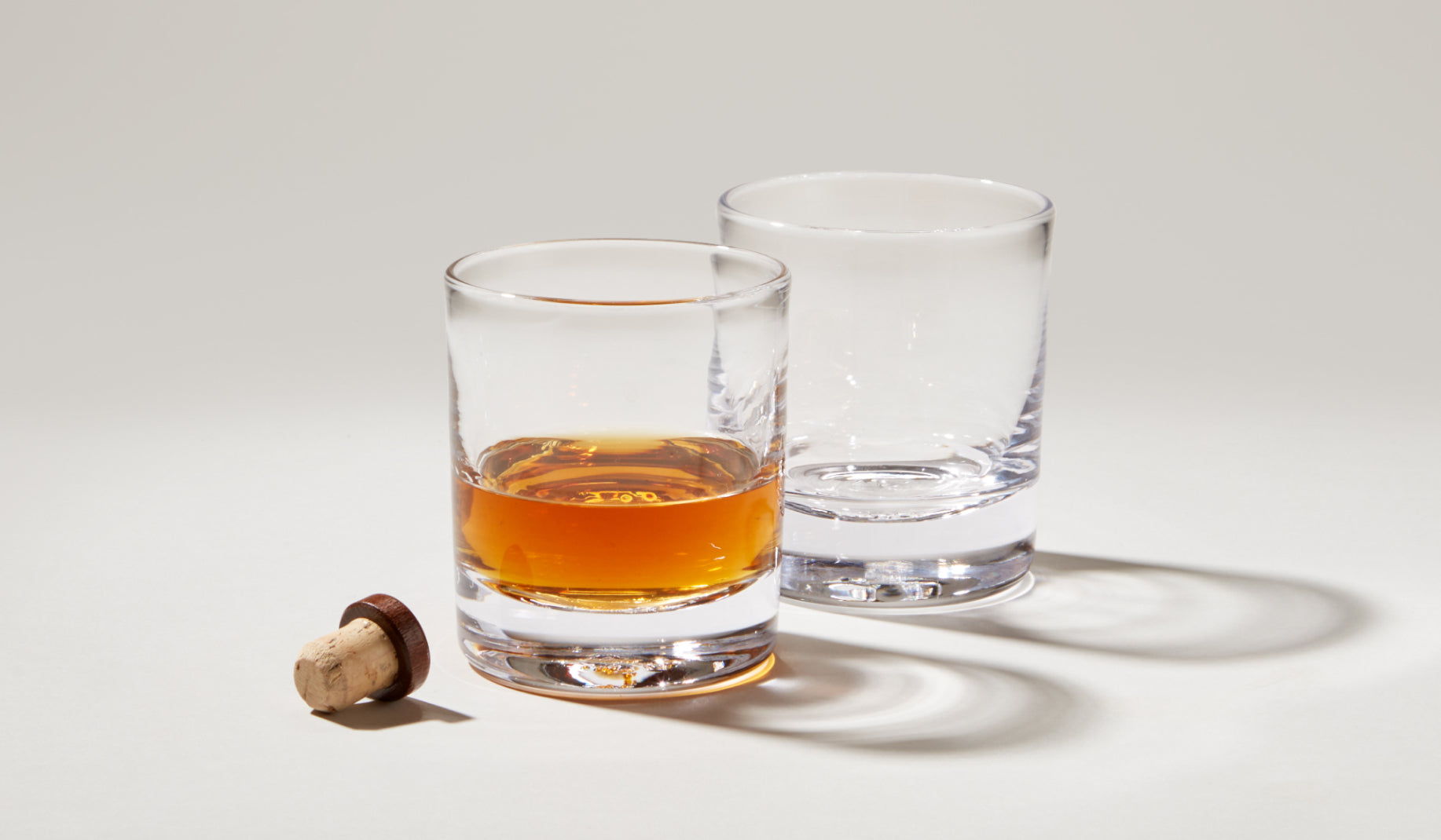 Two Simon Pearce Ascutney double old fashioned glasses on a neutral background, one filled with amber whiskey and a cork stopper alongside