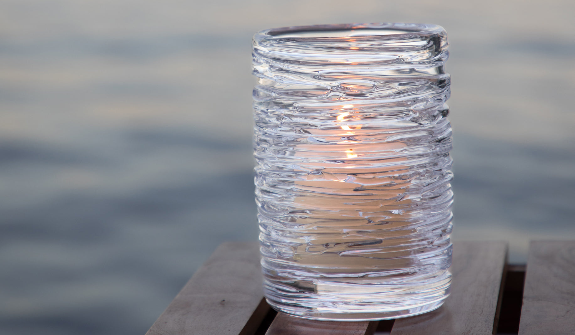 A Simon Pearce Echo Lake hurricane candleholder with a ribbed texture holding a lit pillar candle, displayed on a wood dock with calm water in the background
