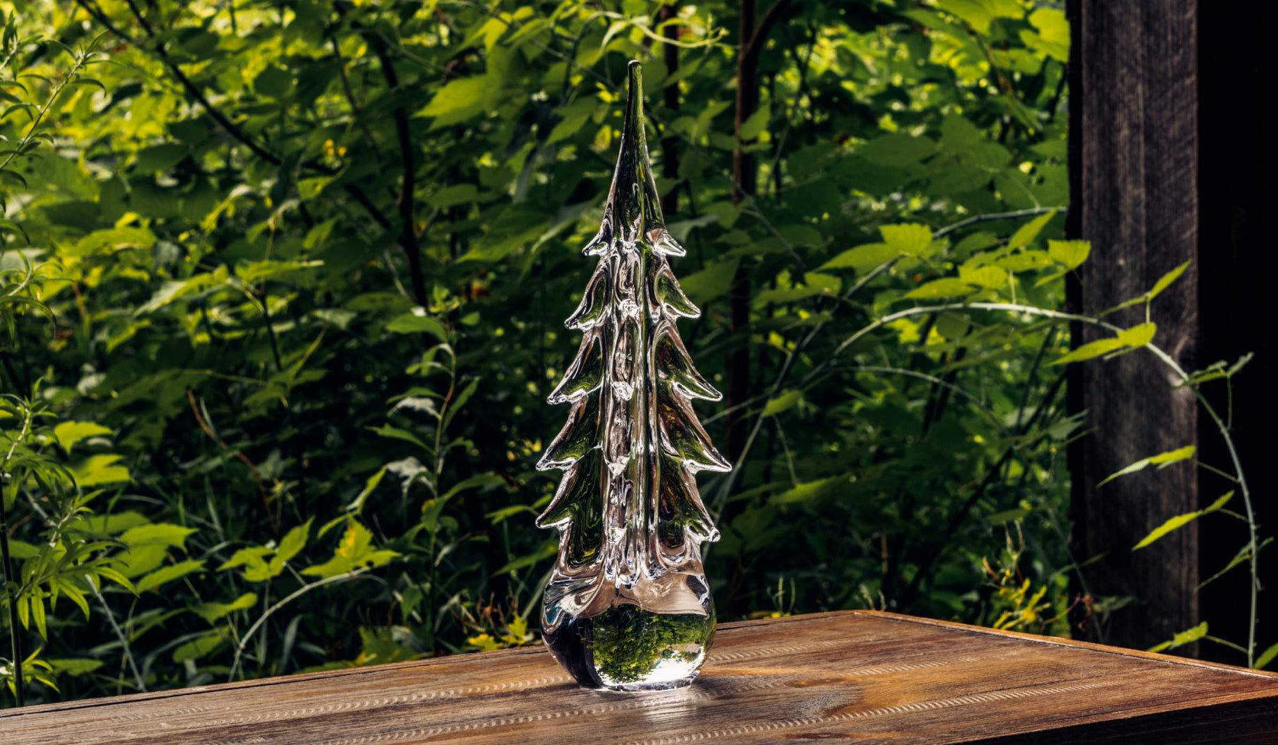 Simon Pearce Five Sided Evergreen handblown glass tree sculpture displayed on a wood surface outdoors against lush green foliage