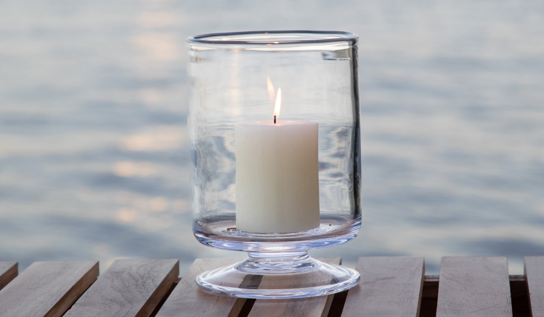 A Simon Pearce Nantucket hurricane candleholder with a lit pillar candle displayed on a wood dock with calm water reflecting the sky in the background