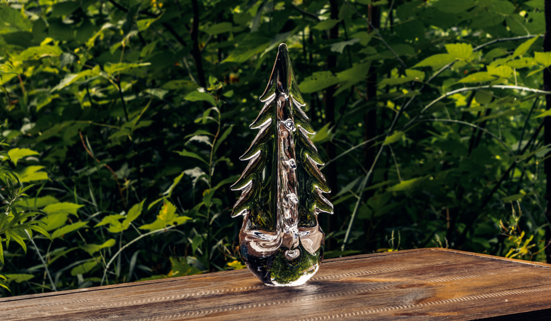 Simon Pearce Vermont Evergreen handblown glass tree sculpture on a wood surface outdoors, reflecting the surrounding forest foliage