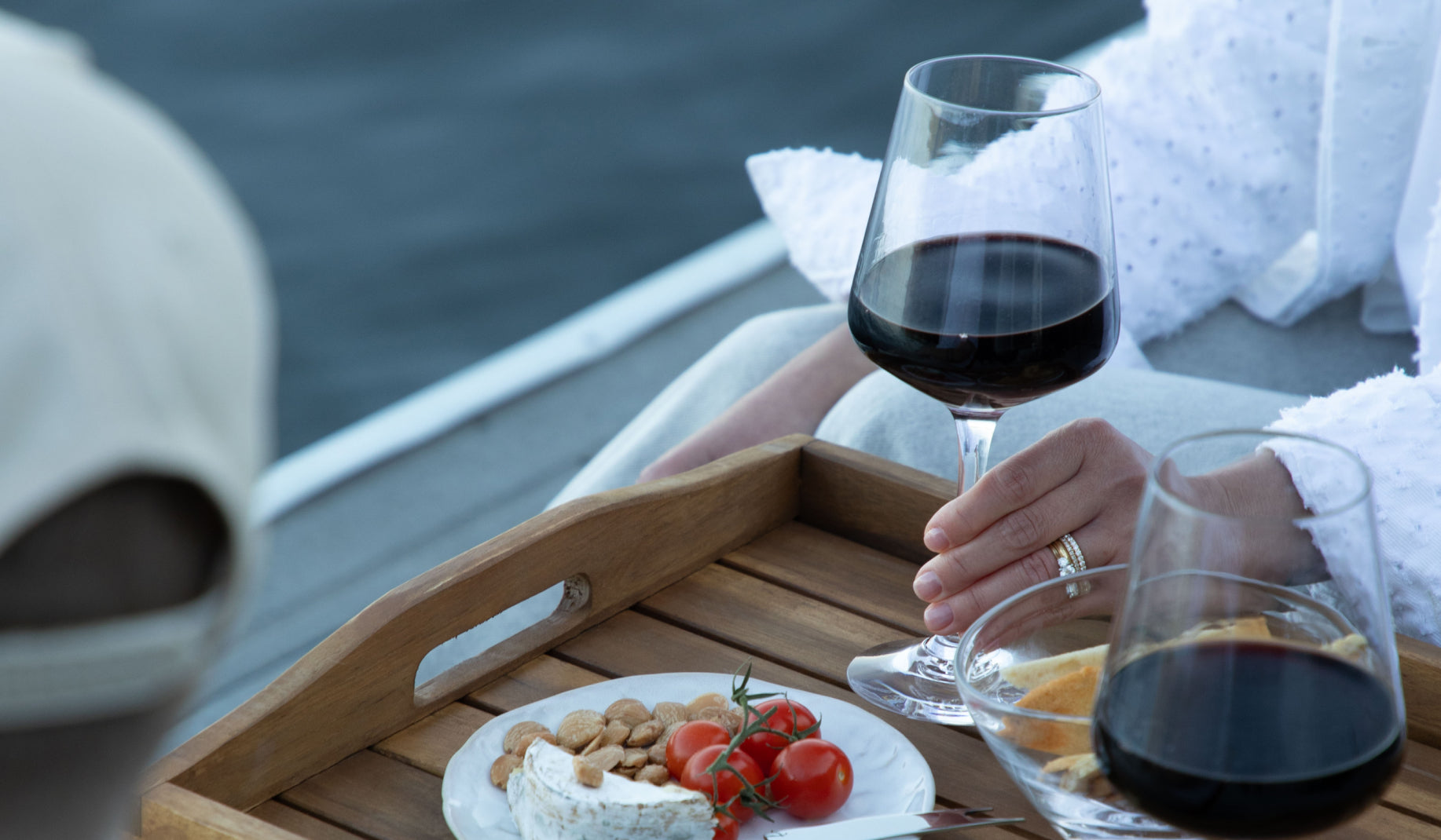 A hand holding a Simon Pearce Vintner red wine glass on an outdoor wood tray beside a second wine glass, cheese board, and cherry tomatoes with water in the background