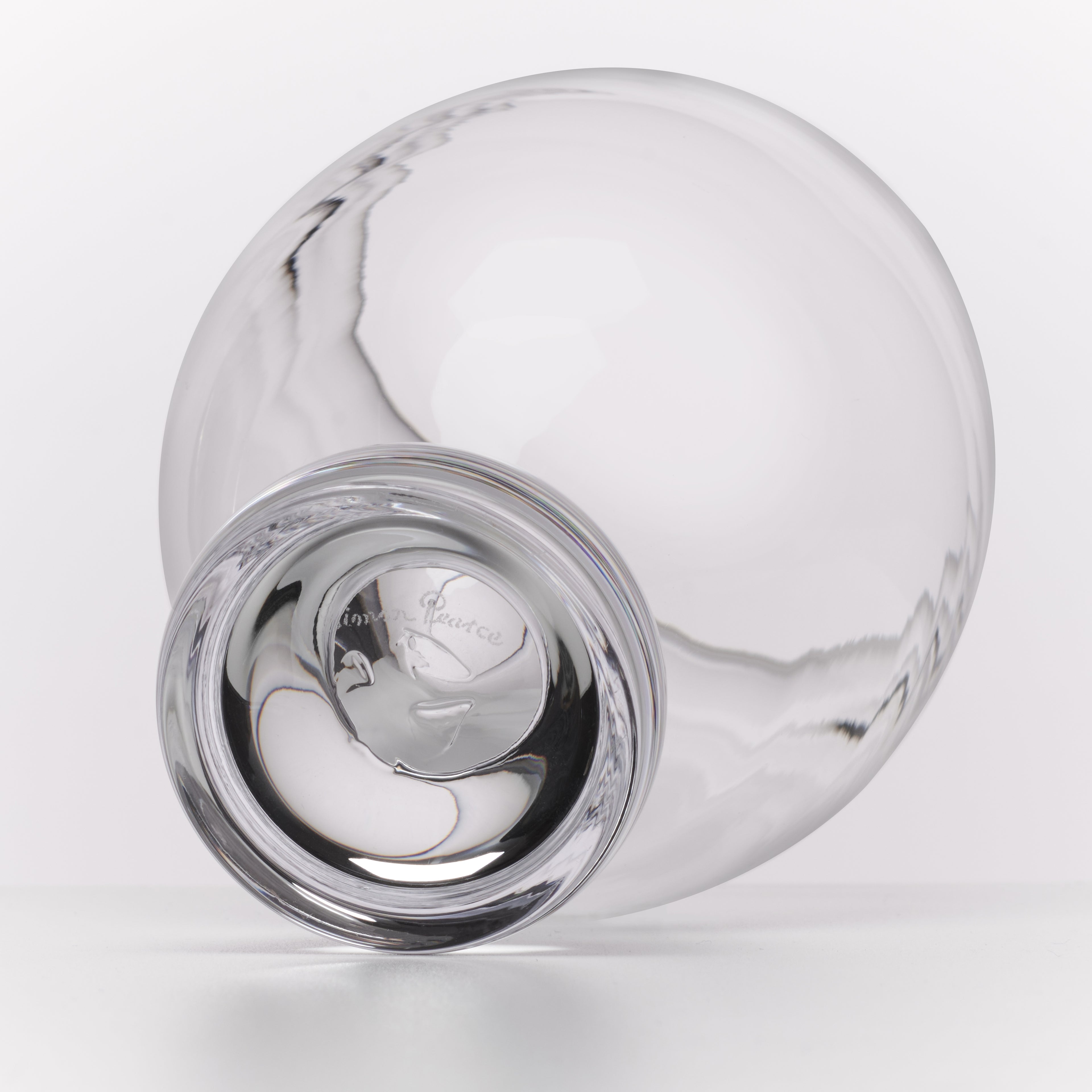 A clear glass carafe with a wide rounded body and a short, thick base, photographed on a clean white background. The outside of the base shows a printed logo.