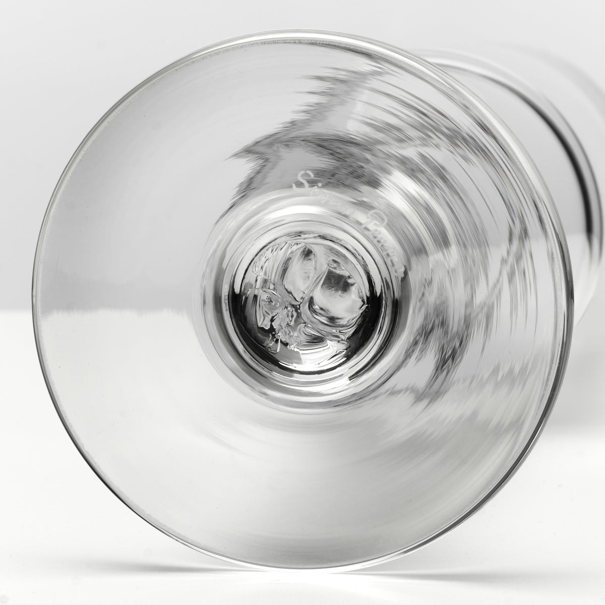 A close-up studio shot of a clear glass wine glass viewed from the bottom, showing the circular base and subtle reflections on the transparent surface against a light gray background.