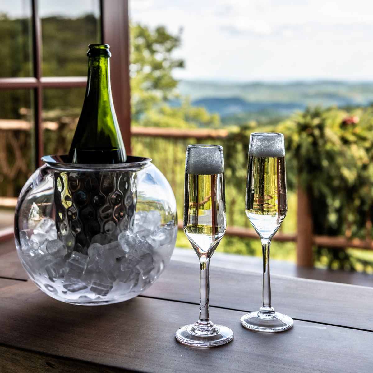 Two clear flute glasses filled with champagne displayed next to an ice bucket with a bottle, overlooking a landscape.
