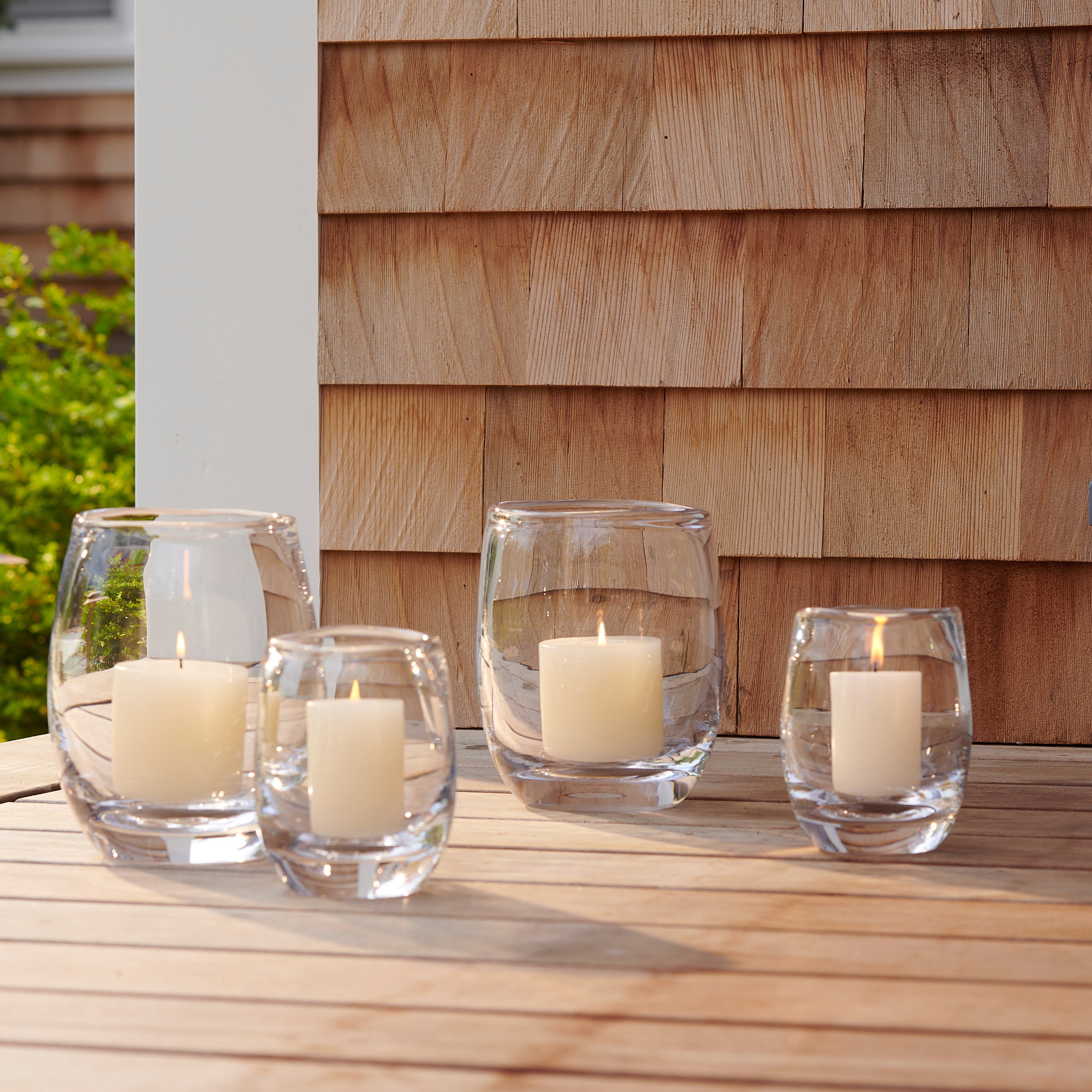Four clear glass votive holders with cream-colored pillar candles arranged on wooden deck.