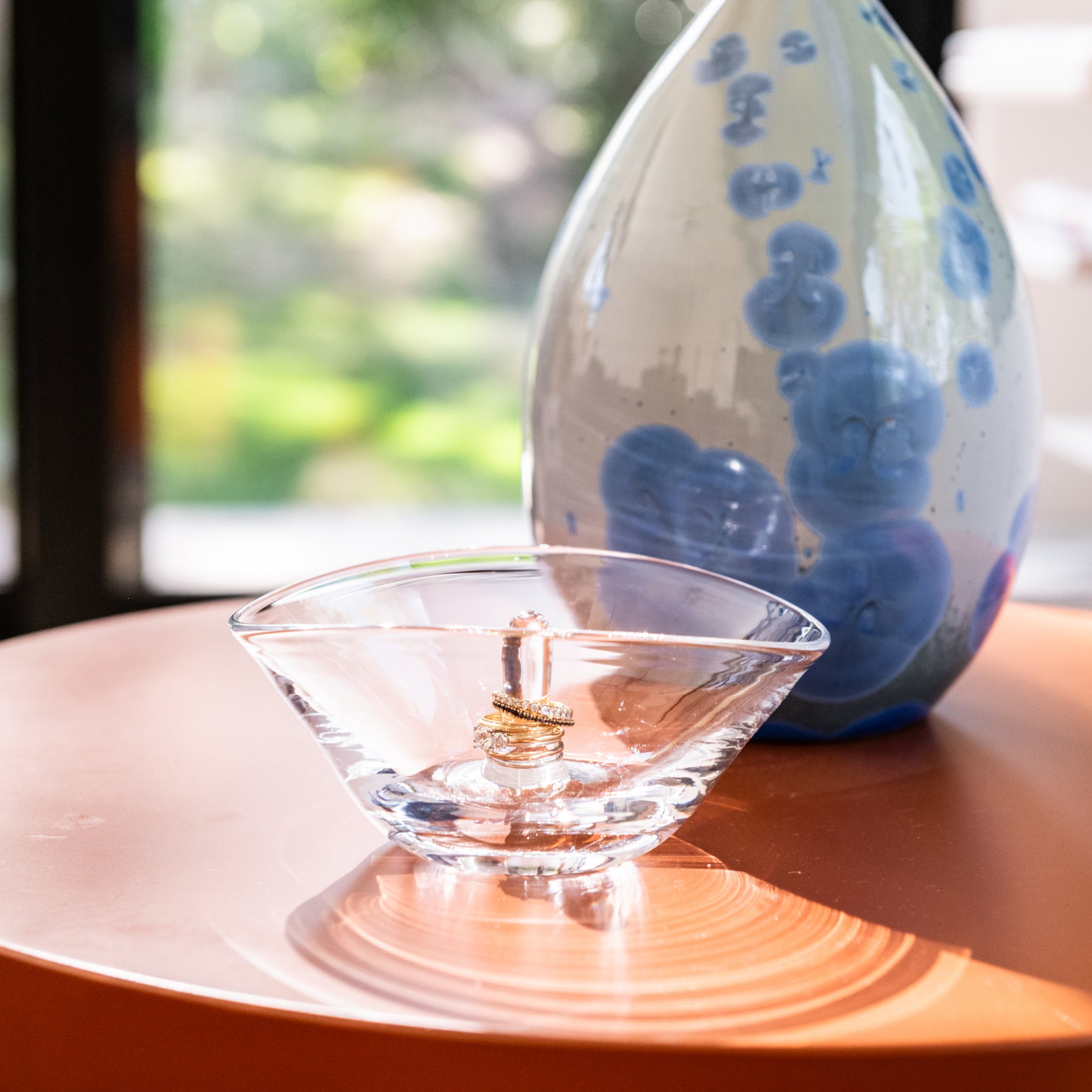 Clear glass ring holder with gold band, displayed with blue and white ceramic vessel in background.