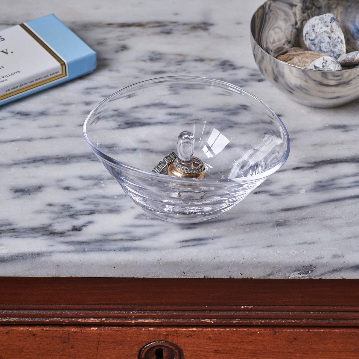 Clear glass bowl ring holder with decorative metal post, displayed on marble surface.
