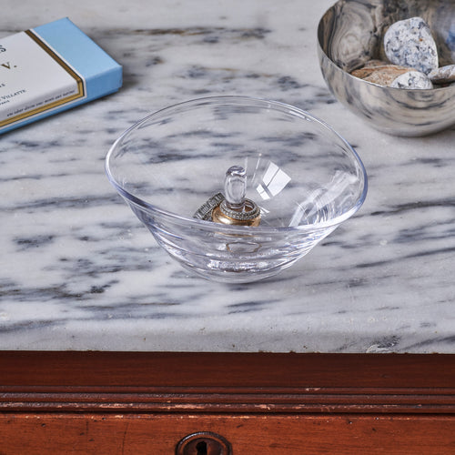 Clear glass bowl ring holder with decorative metal post, displayed on marble surface.