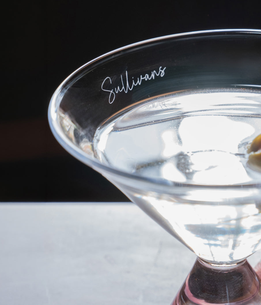Close-up of a Simon Pearce martini glass engraved with "Sullivans" against a dark background