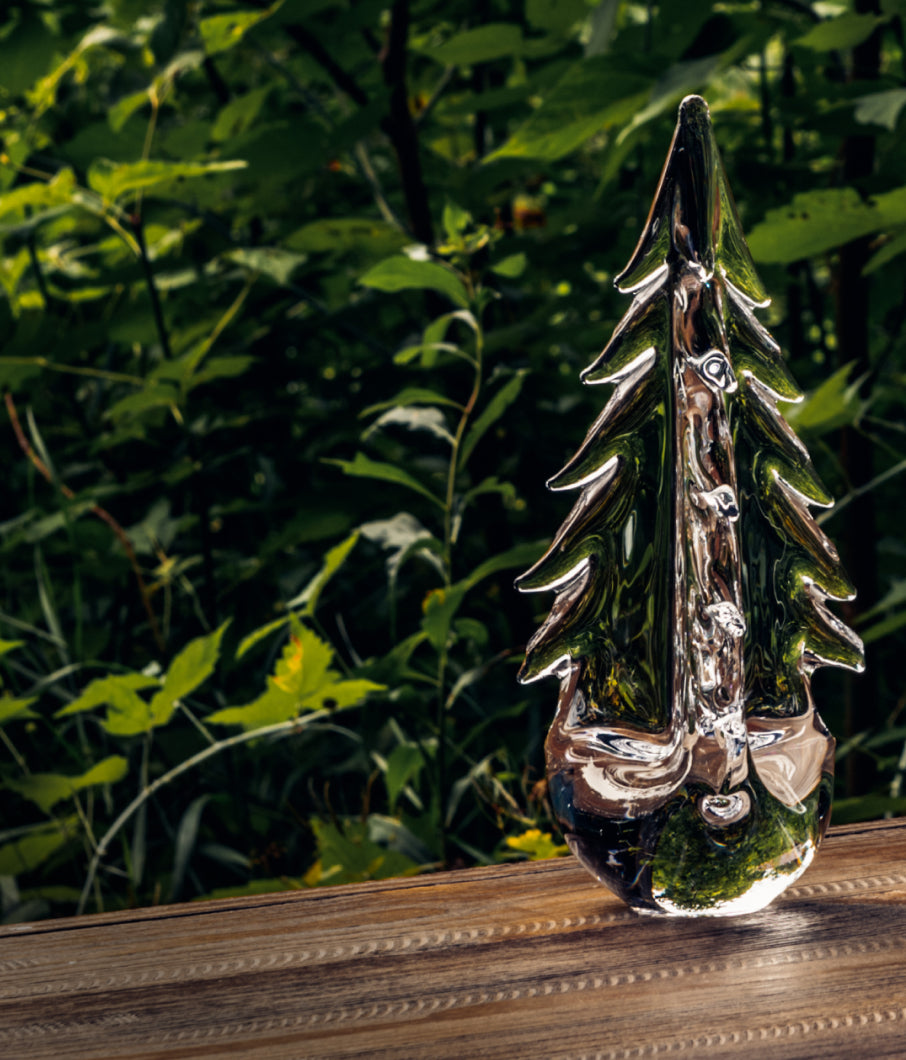 Simon Pearce handblown glass evergreen tree sculpture displayed outdoors on a wooden surface with lush foliage in the background