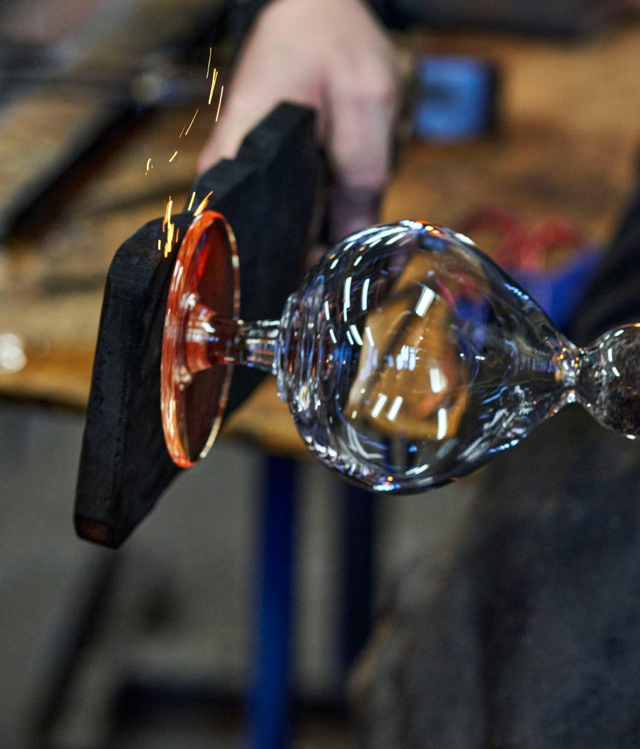 A Simon Pearce artisan shaping a glowing hot handblown wine glass with a wooden paddle, sparks flying in the studio