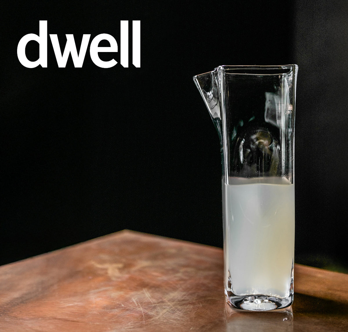 Dwell magazine feature: Simon Pearce handblown glass pitcher with angled lip, filled with a milky white liquid, on a worn copper surface against a dark background