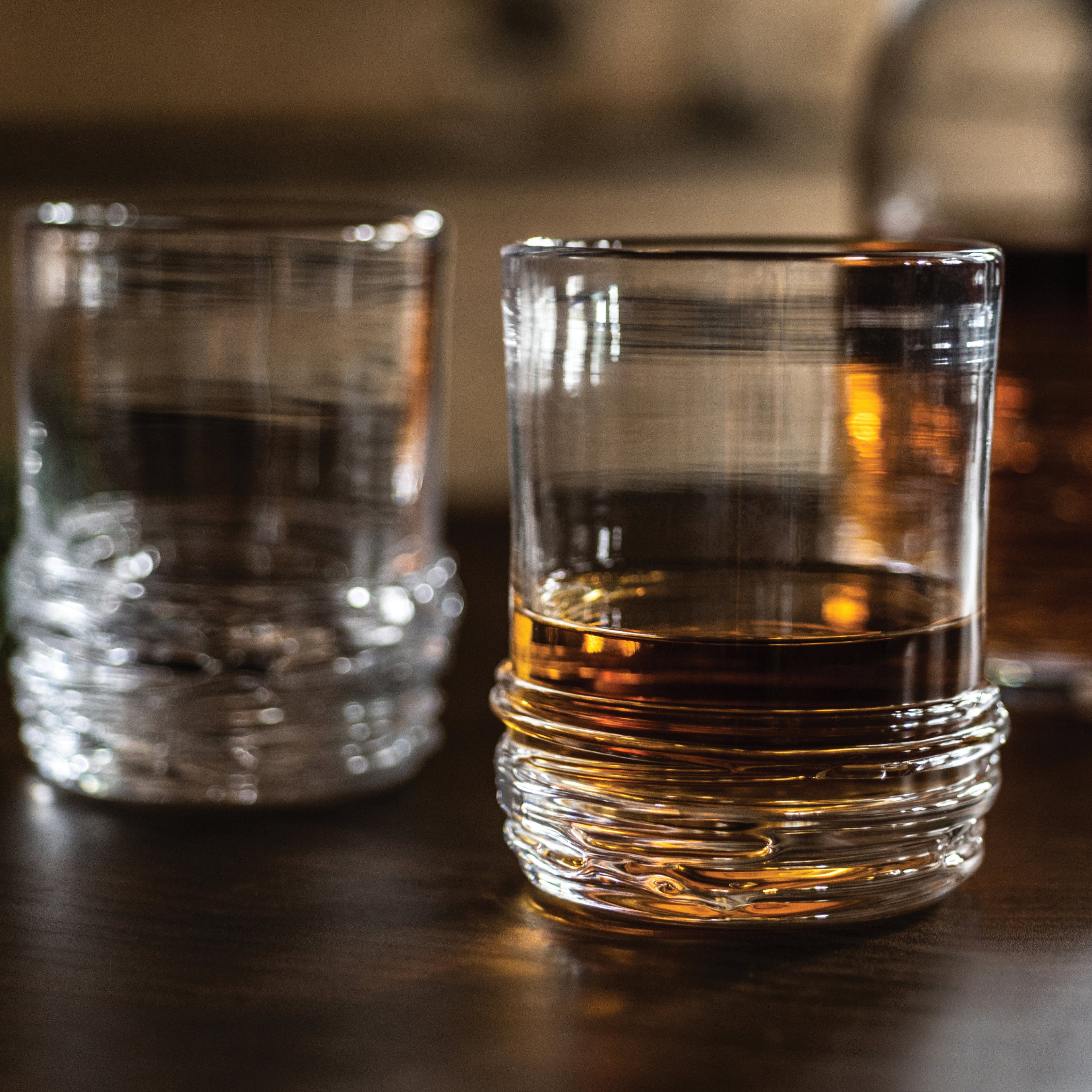 Two crystal rocks glasses with textured bases, one containing amber whiskey and one empty, on a dark surface.