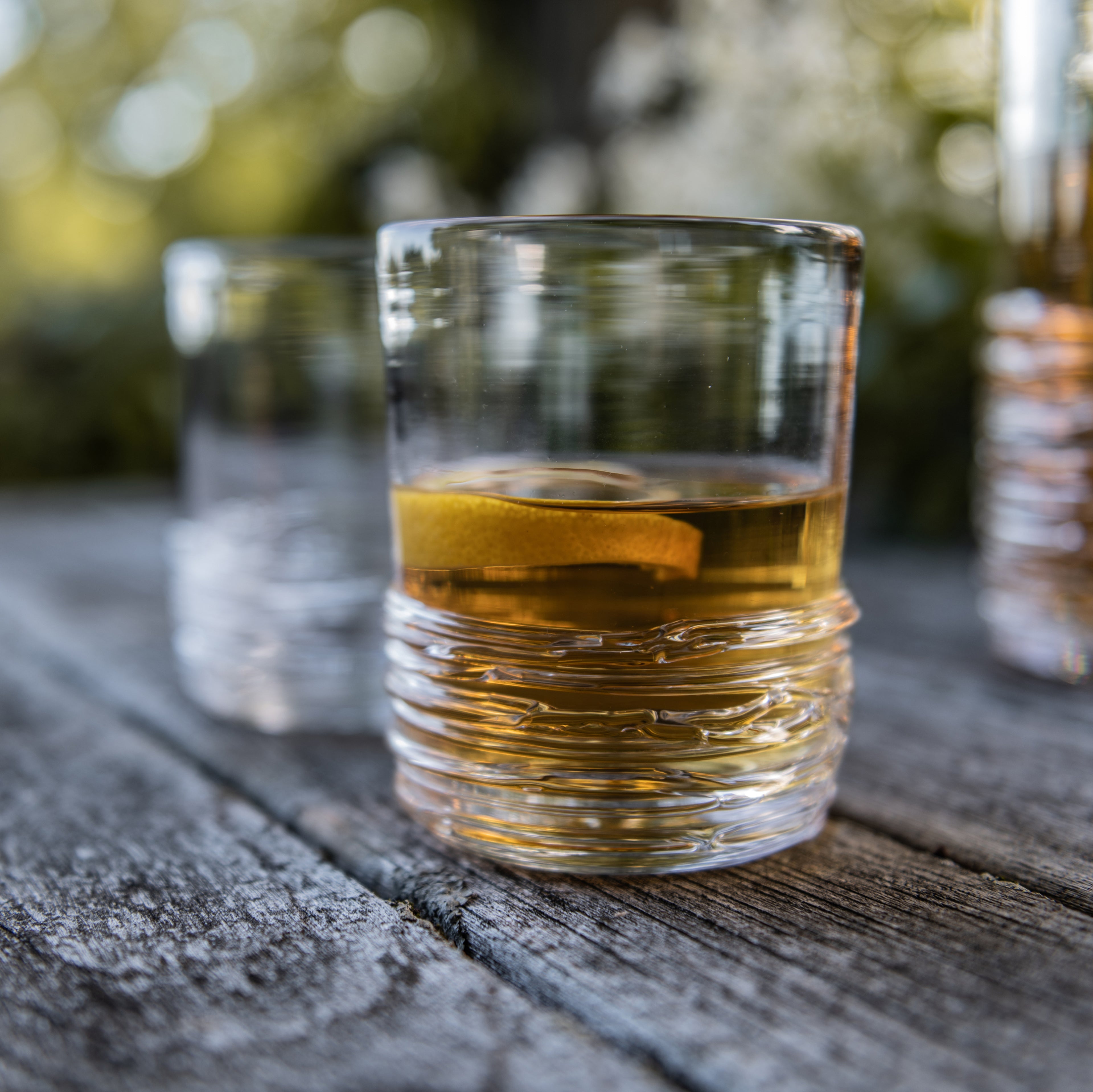 Glass of amber whiskey with ice on wooden surface, additional glasses visible in background.