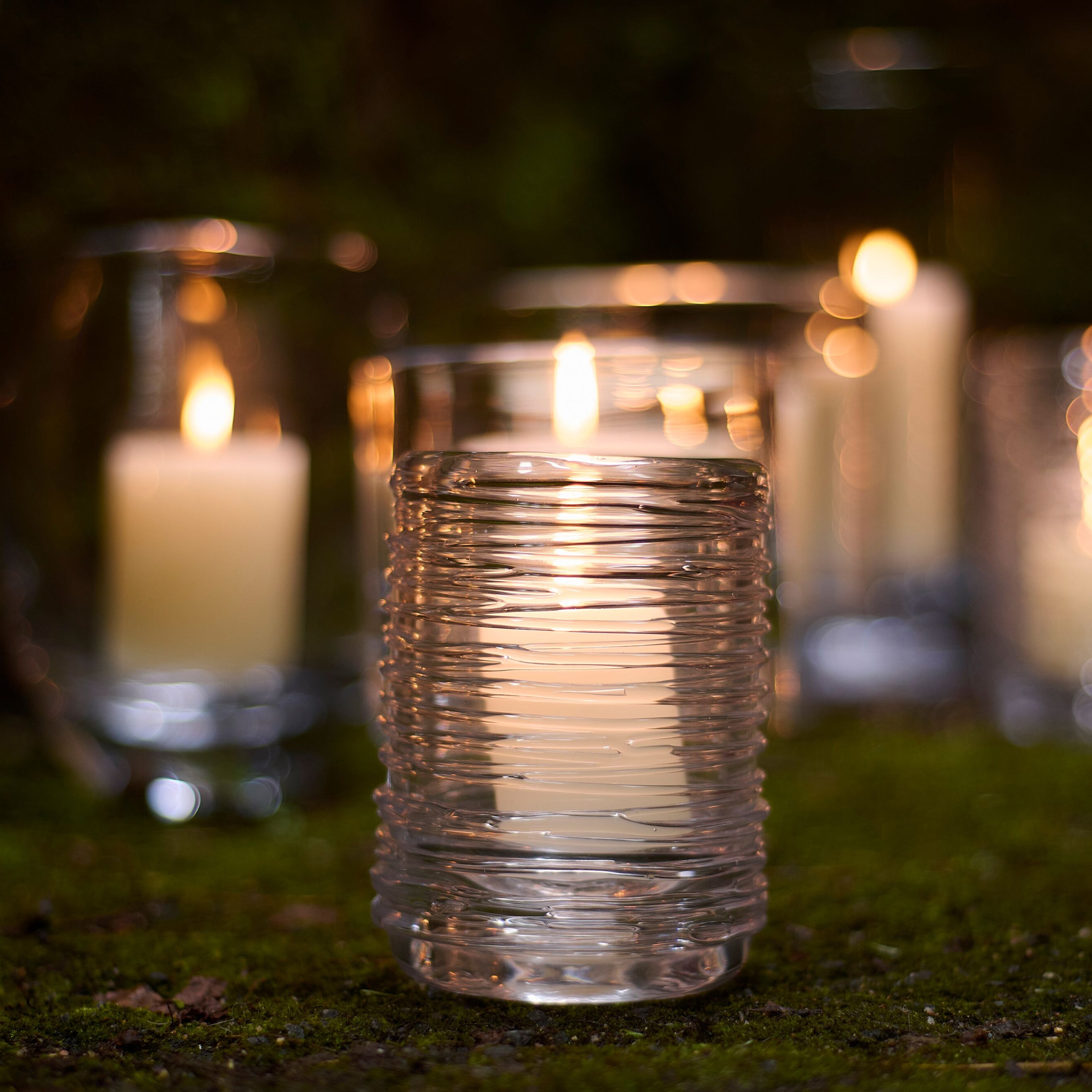 Multiple clear glass hurricane candles with textured surfaces lit and arranged outdoors on grass at dusk.