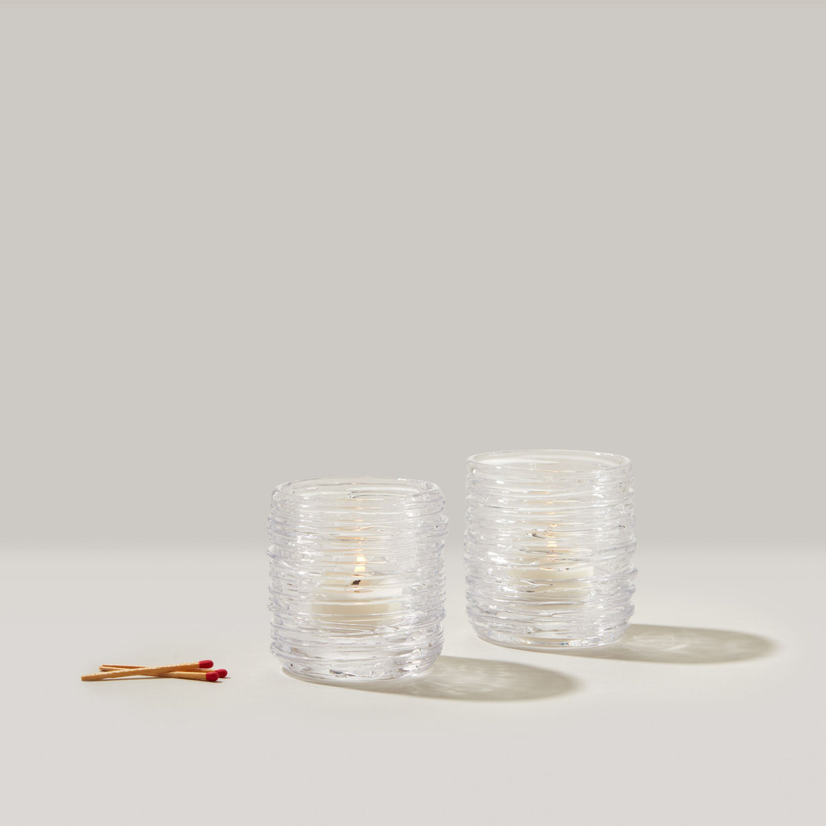 Two clear glass tealight candles with cream-colored wax and two wooden matches with red tips on neutral background.