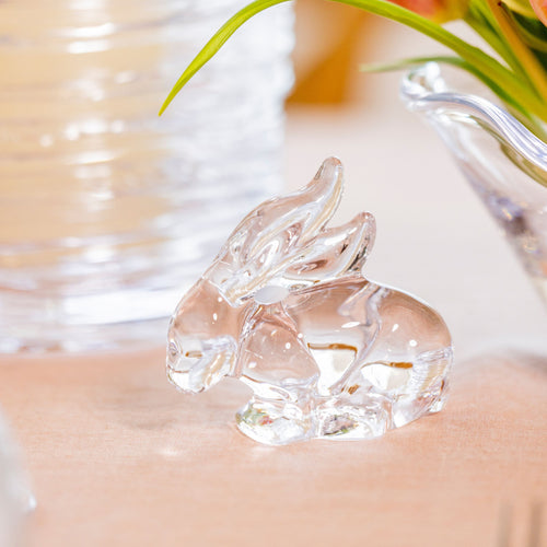 Clear glass rabbit figurine with raised ears on a beige surface near potted plant.