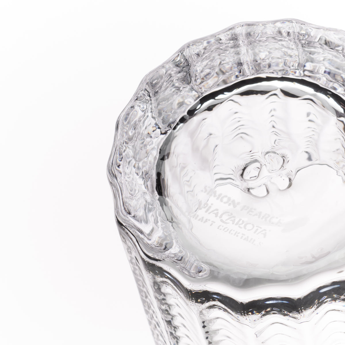 A close-up of a clear textured glass tumbler, showing the inside base engraved with 'Simon Pearce Via Carota Craft Cocktails' on a white background.