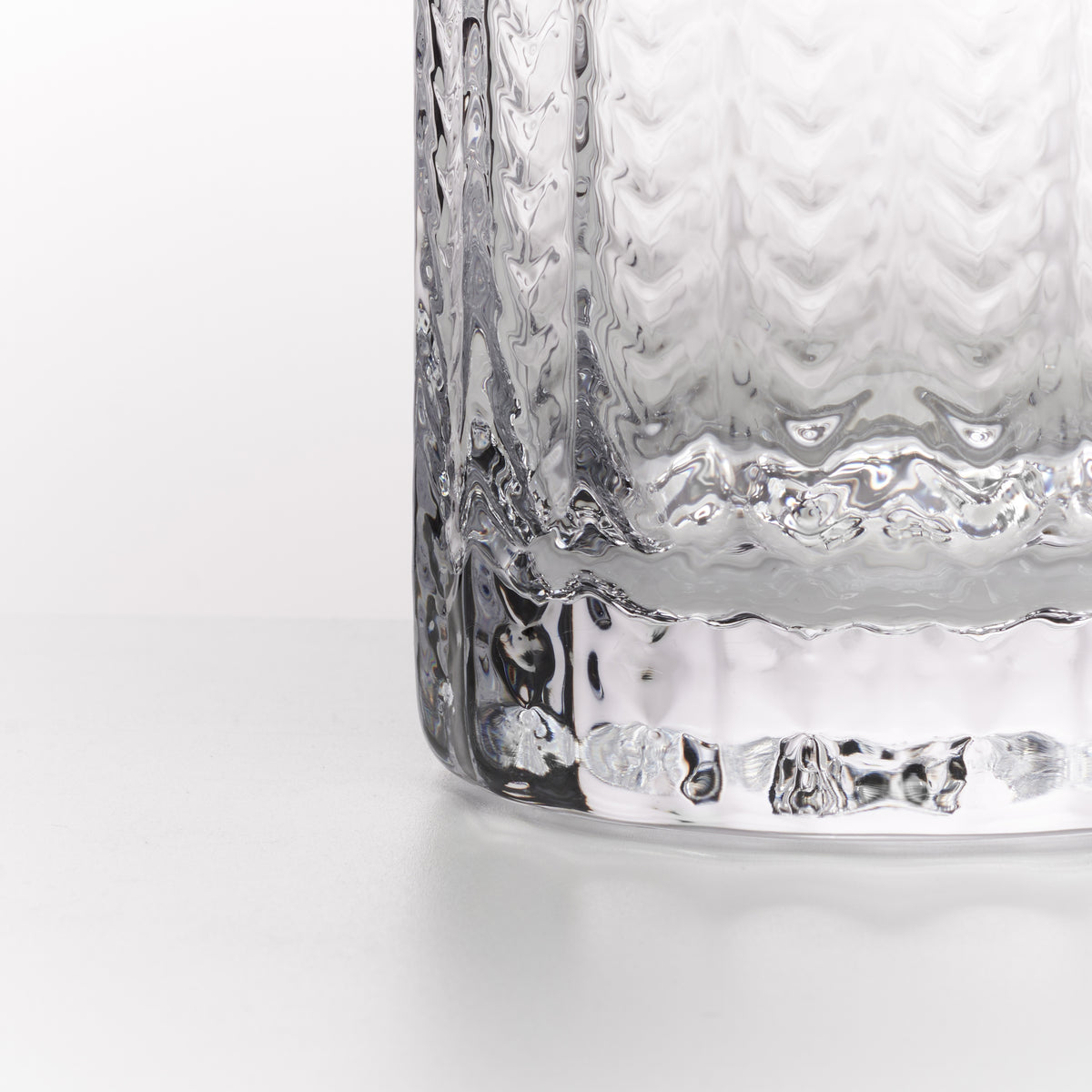 A close-up of a clear textured glass tumbler with a wavy, hammered pattern and a thick ridged base, set against a clean white background.