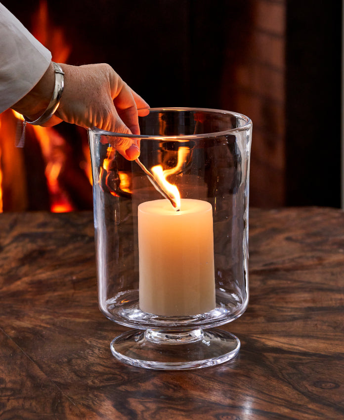 A hand lighting a pillar candle inside a Simon Pearce handblown glass hurricane candleholder on a wood surface with a fireplace glowing in the background