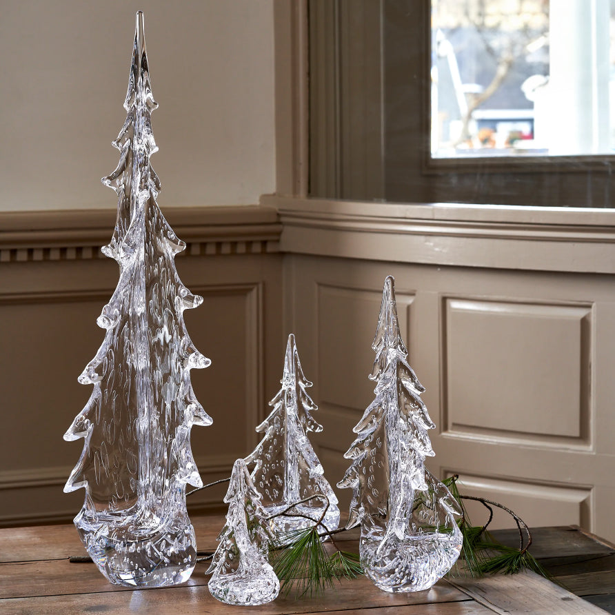 Three Simon Pearce handblown clear glass evergreen tree sculptures in varying sizes displayed on a wood floor beside a window with pine branch accents