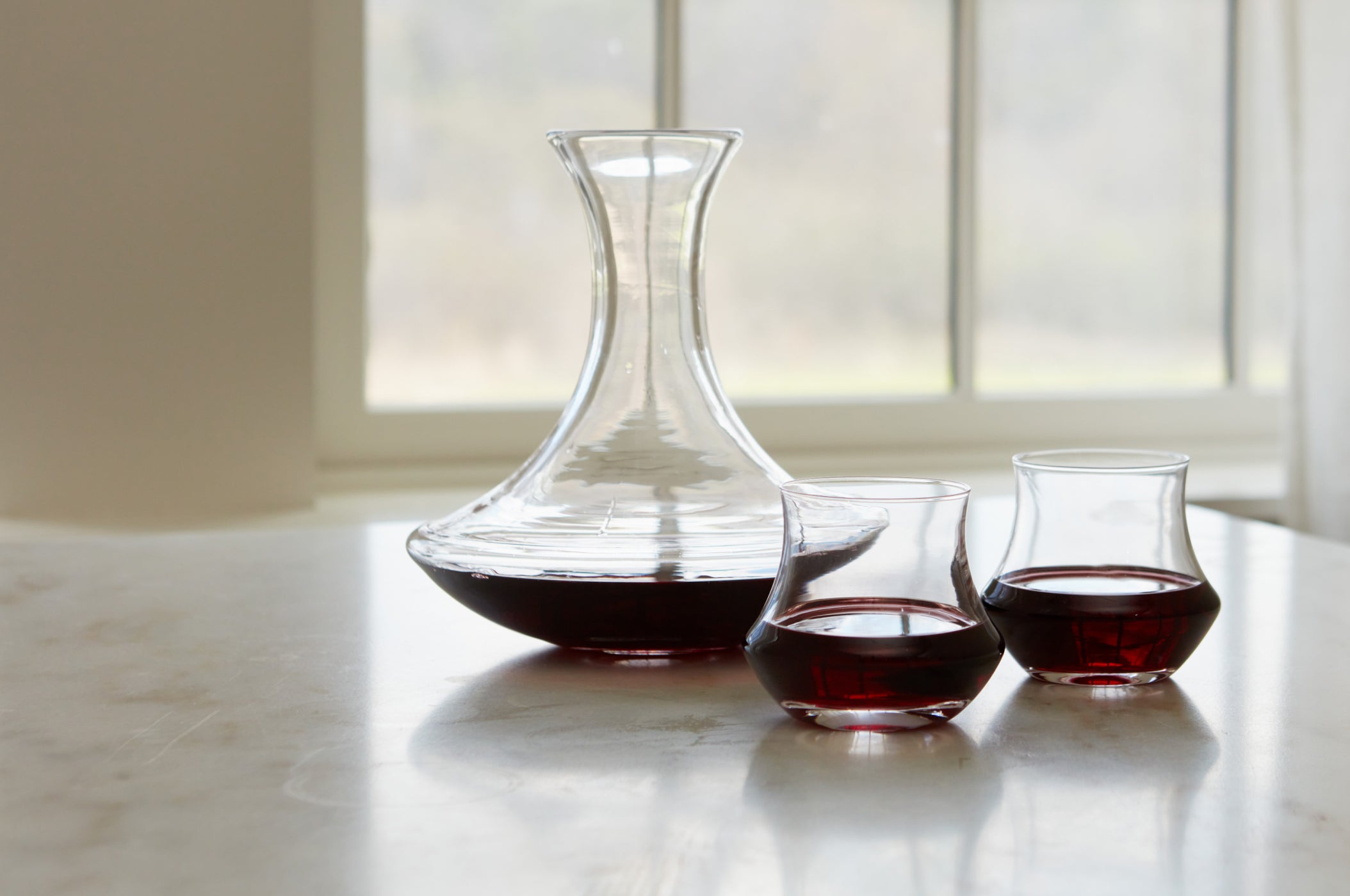 A Simon Pearce handblown glass wine decanter filled with red wine beside two short tumbler glasses on a marble surface in front of a bright window