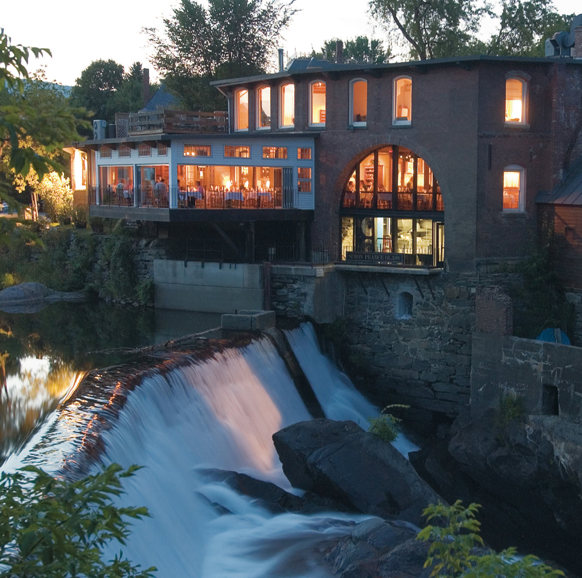 The Simon Pearce Mill restaurant and glassblowing studio in Quechee, Vermont, illuminated at dusk above a rushing waterfall on the Ottauquechee River