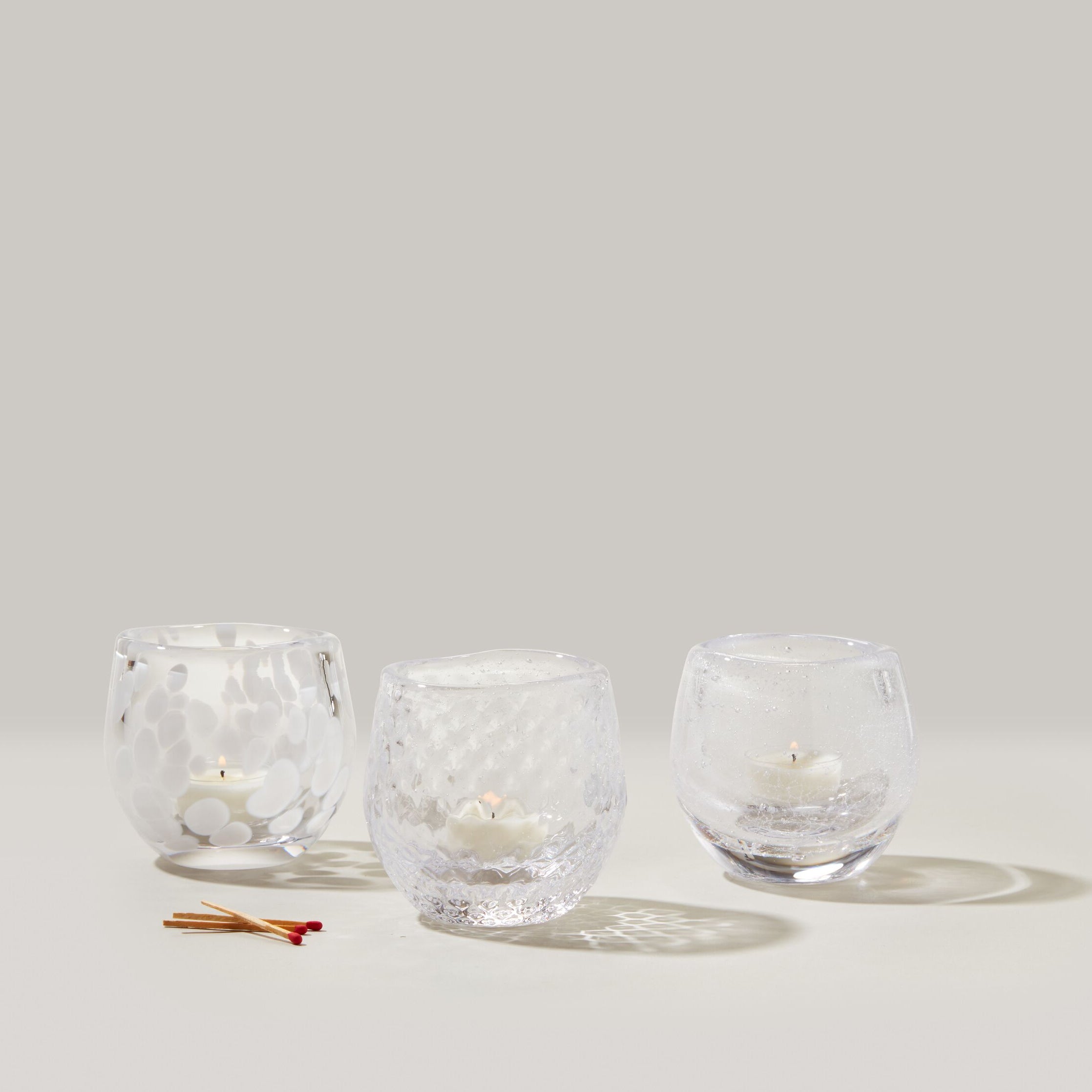 Three clear glass tealight holders with textured surfaces containing white tealight candles, displayed with matches.