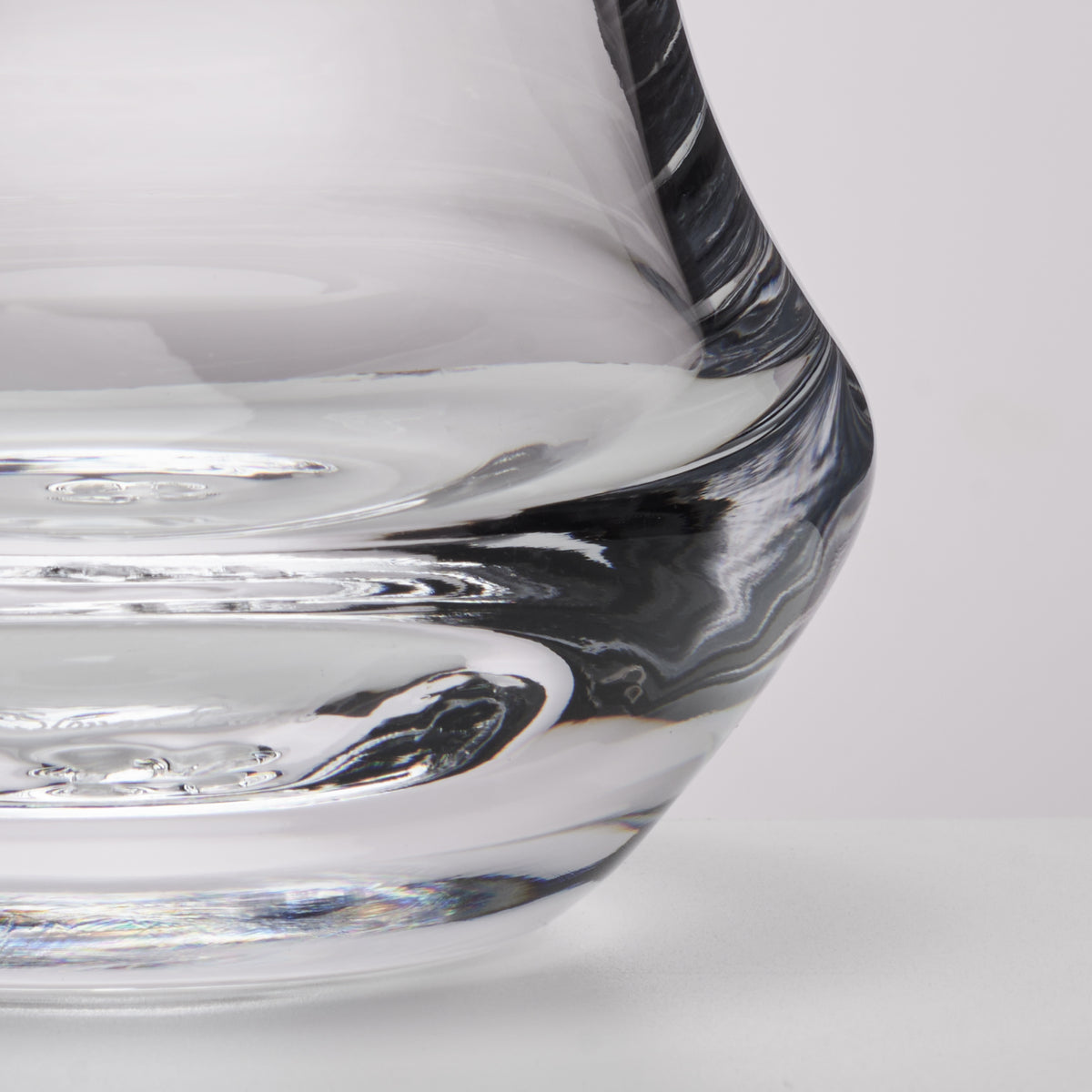 Close-up of a clear glass vessel with a curved, sculptural rim showing reflective glass textures on a light gray background.