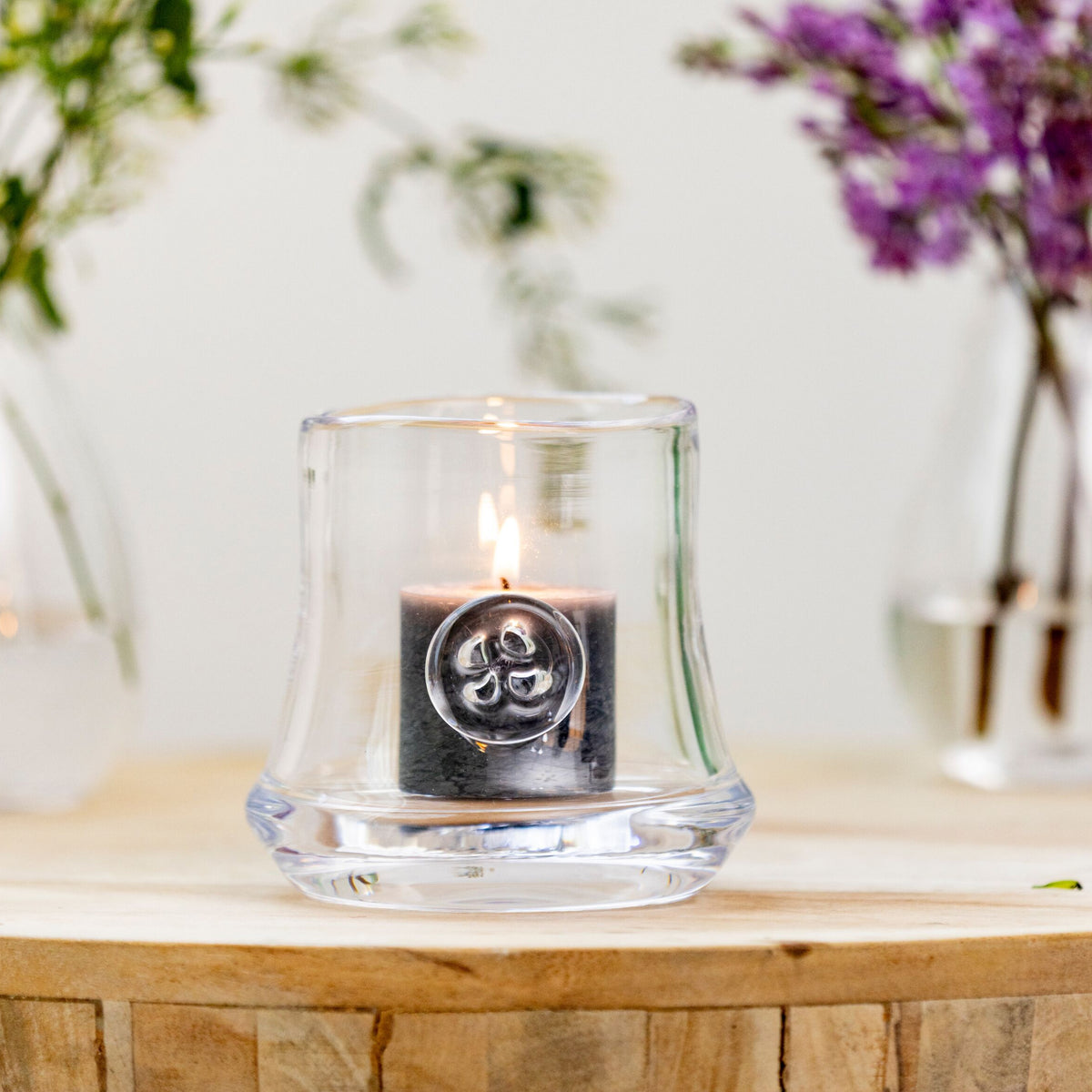 Grey pillar candle with decorative circular medallion, displayed in clear glass holder with lit flame.