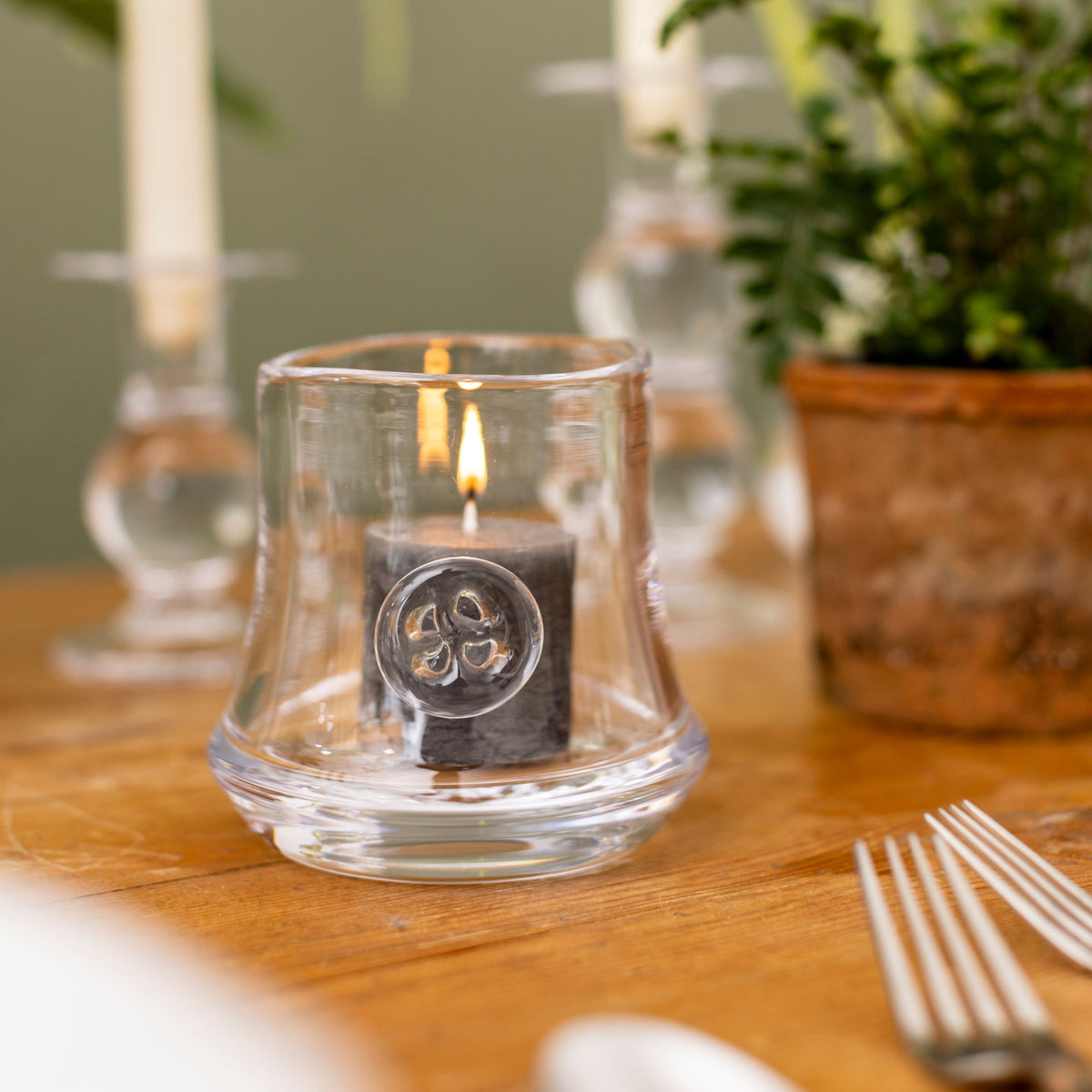 Clear glass hurricane candle holder with lit black candle featuring circular emblem, on wooden surface.