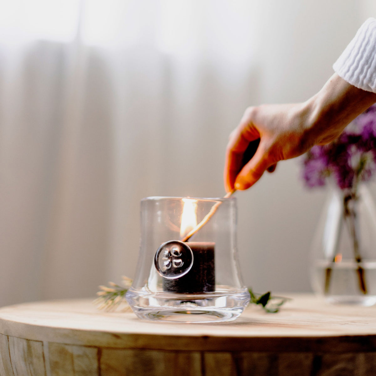 Black candle with decorative medallion inside clear glass holder, hand lighting the wick.