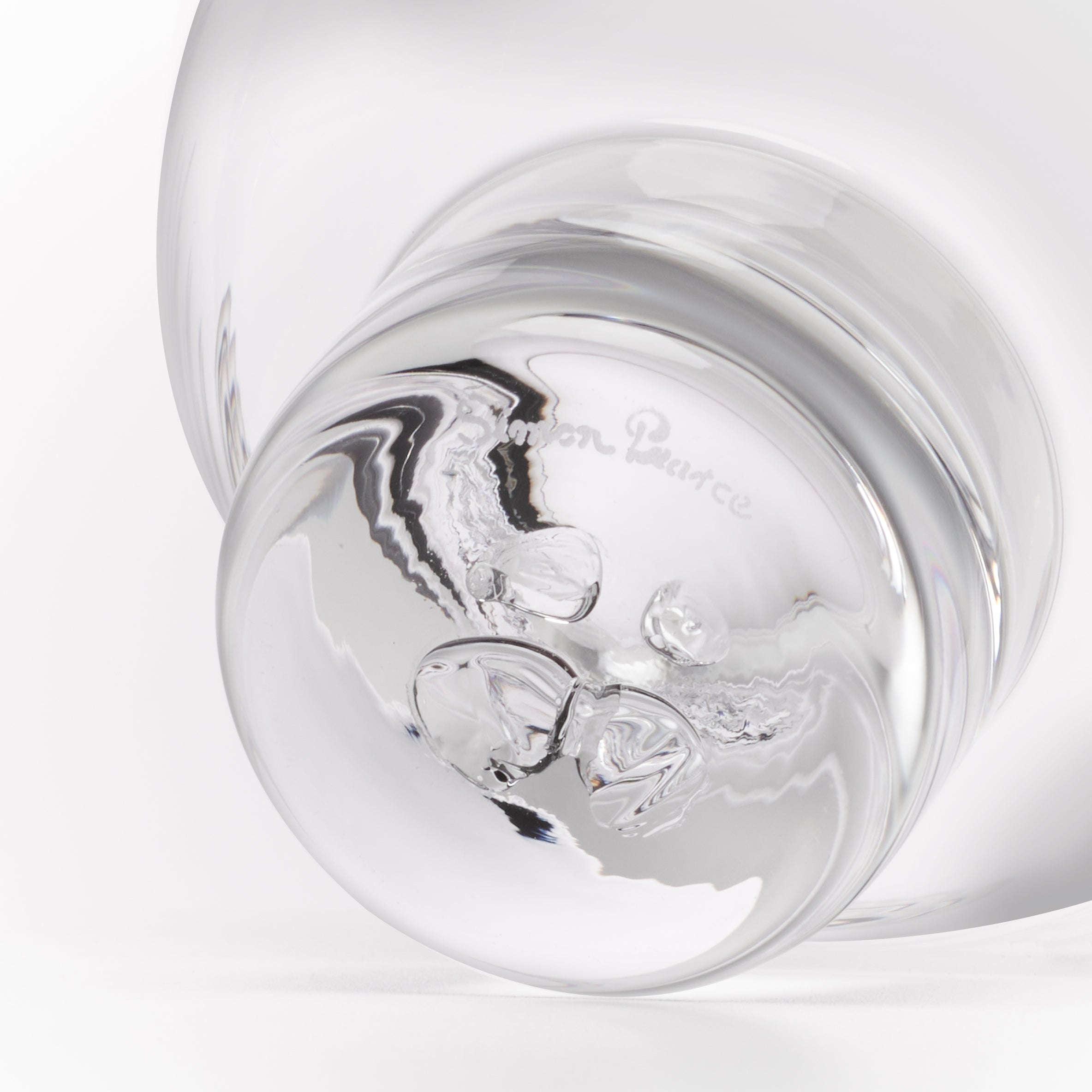 A close-up studio shot of a clear glass object, showing a rounded transparent body and a reflective base with the faint embossed brand name 'Simon Pearce'.