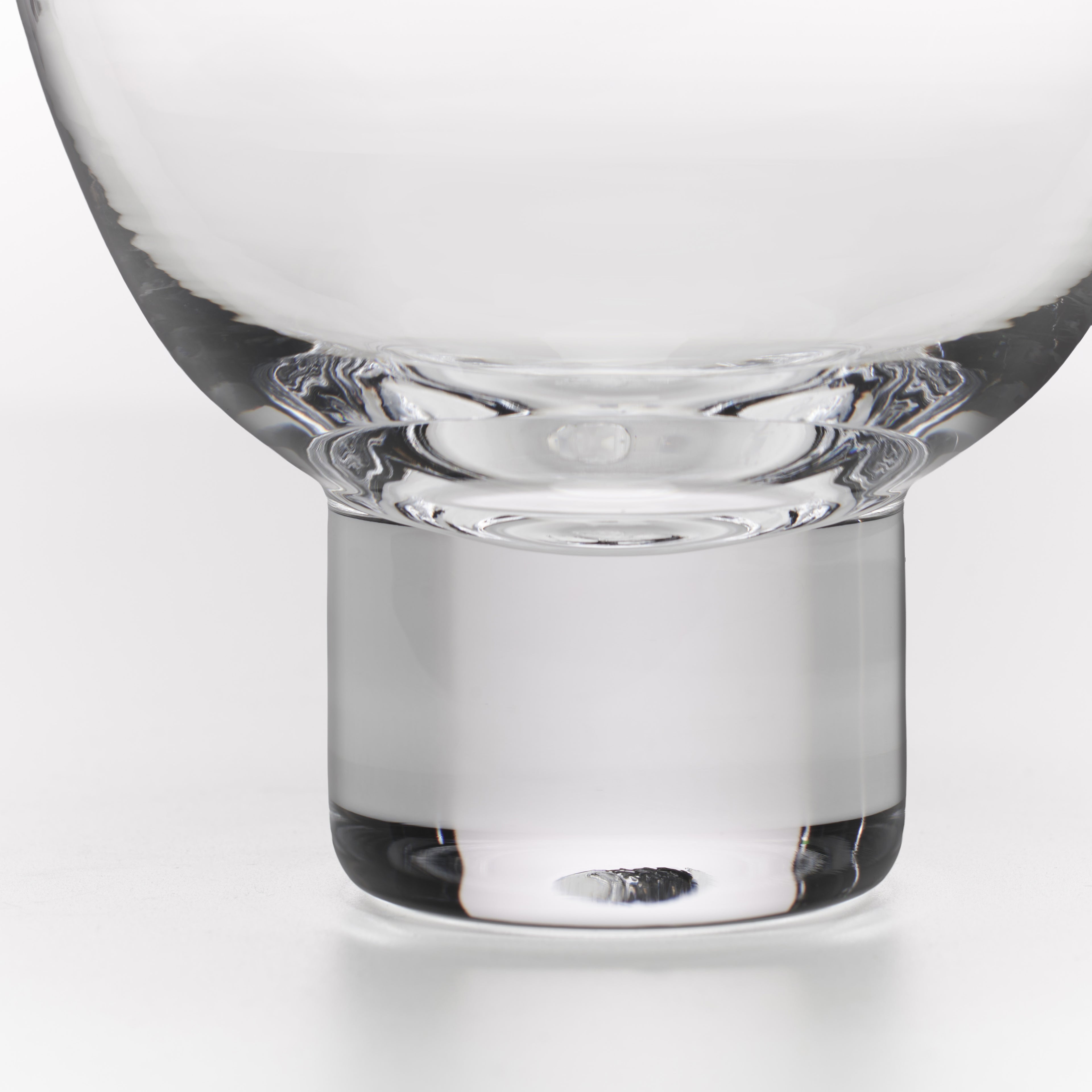 Close-up of a clear glass vessel with a wide, rounded top and a thick cylindrical base, photographed on a light gray studio background.