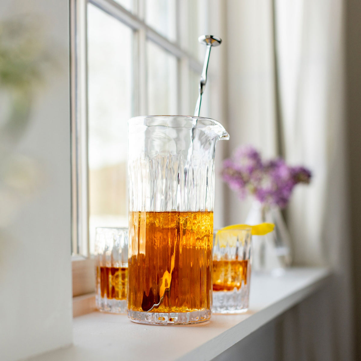 Clear glass pitcher with vertical ribbed texture, filled with amber liquid and bar spoon, displayed with matching glasses.