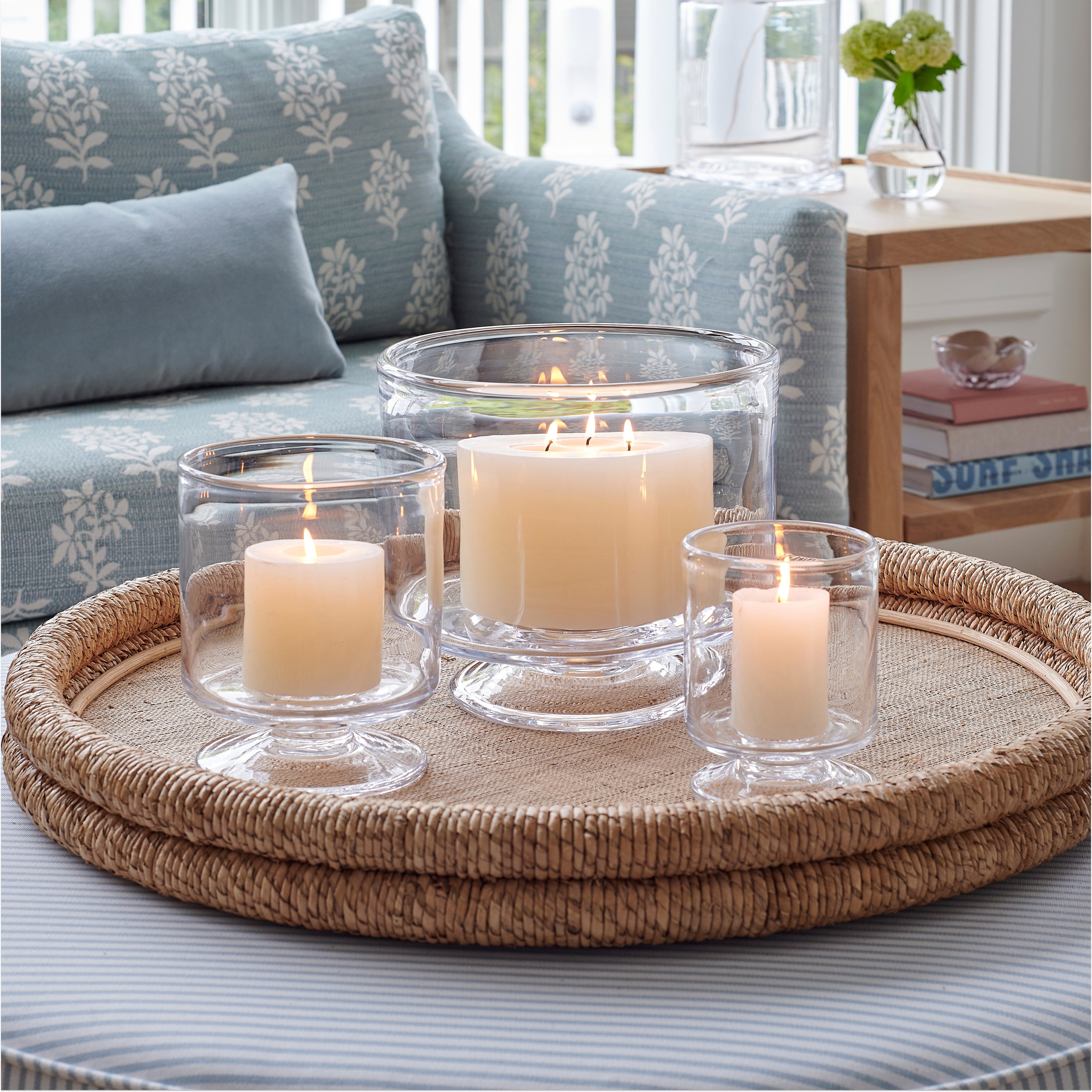 Three cream-colored pillar candles in clear glass hurricane holders arranged on a woven round tray.