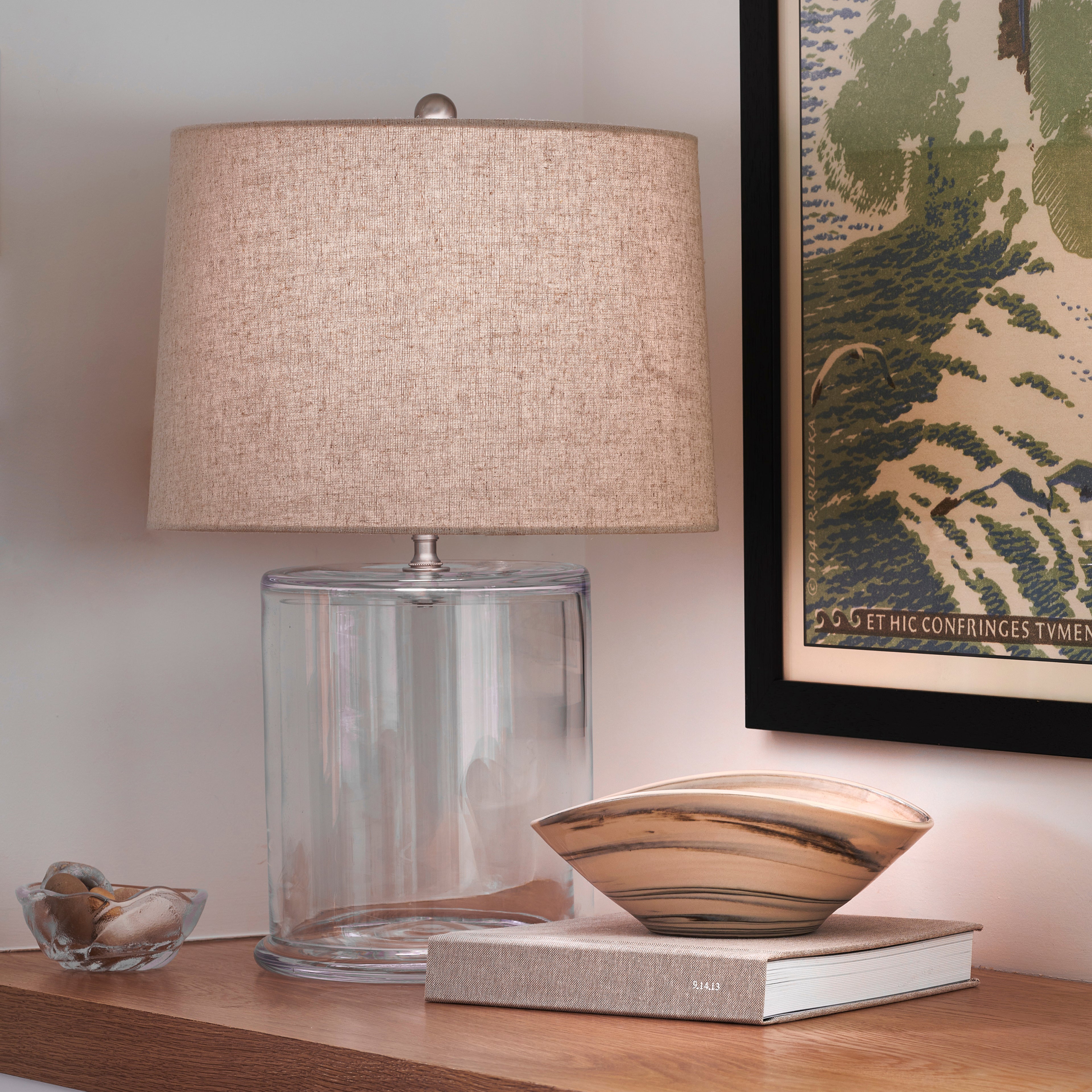 Cylindrical glass lamp base with taupe linen drum shade, displayed on wooden surface with decorative accessories.