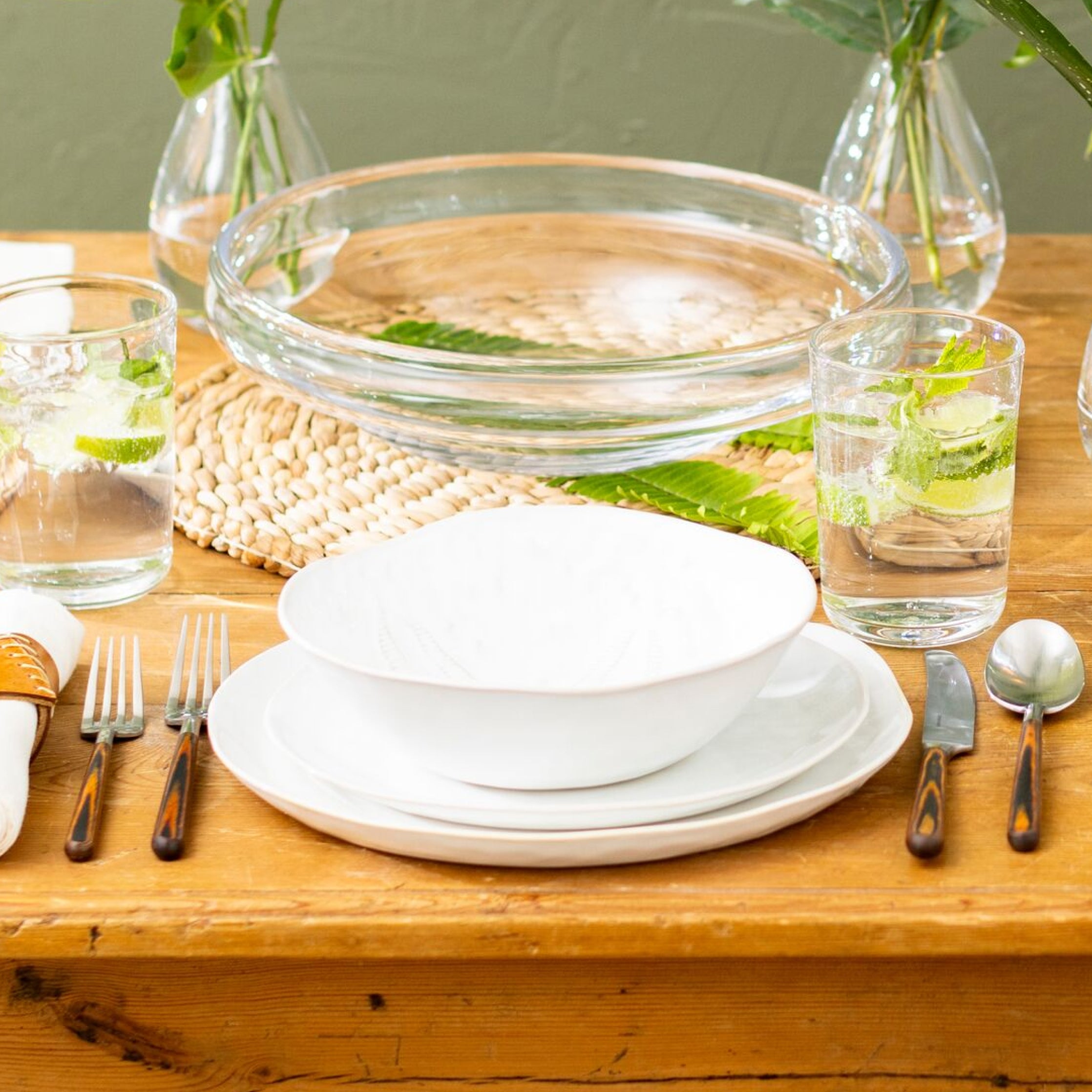 White pasta bowl stacked on white plates on a wooden table with glassware and utensils.