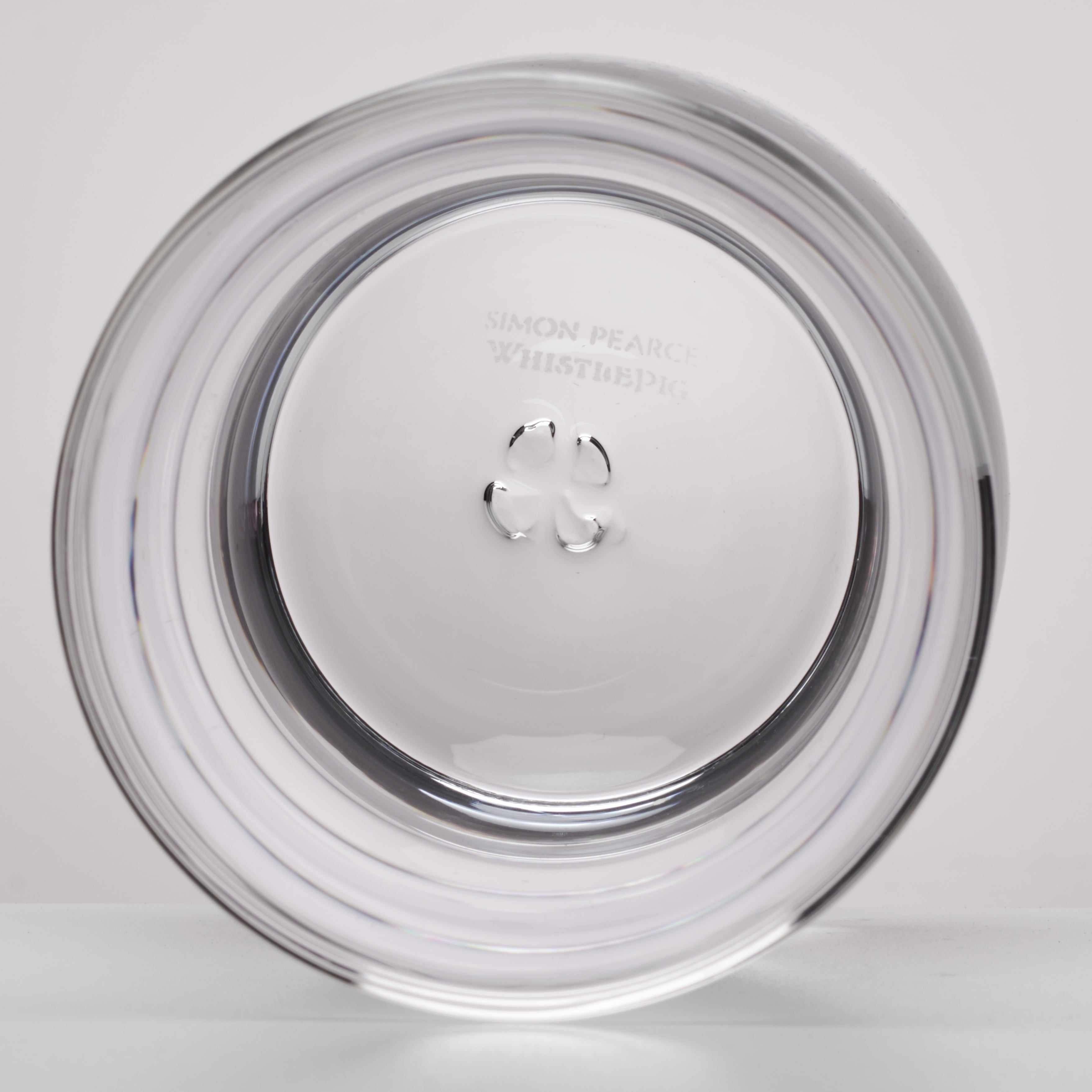 A clear circular glass photographed on a light gray background. Faintly etched text near the top reads 'SIMON PEARCE WHISTLE PIG'.