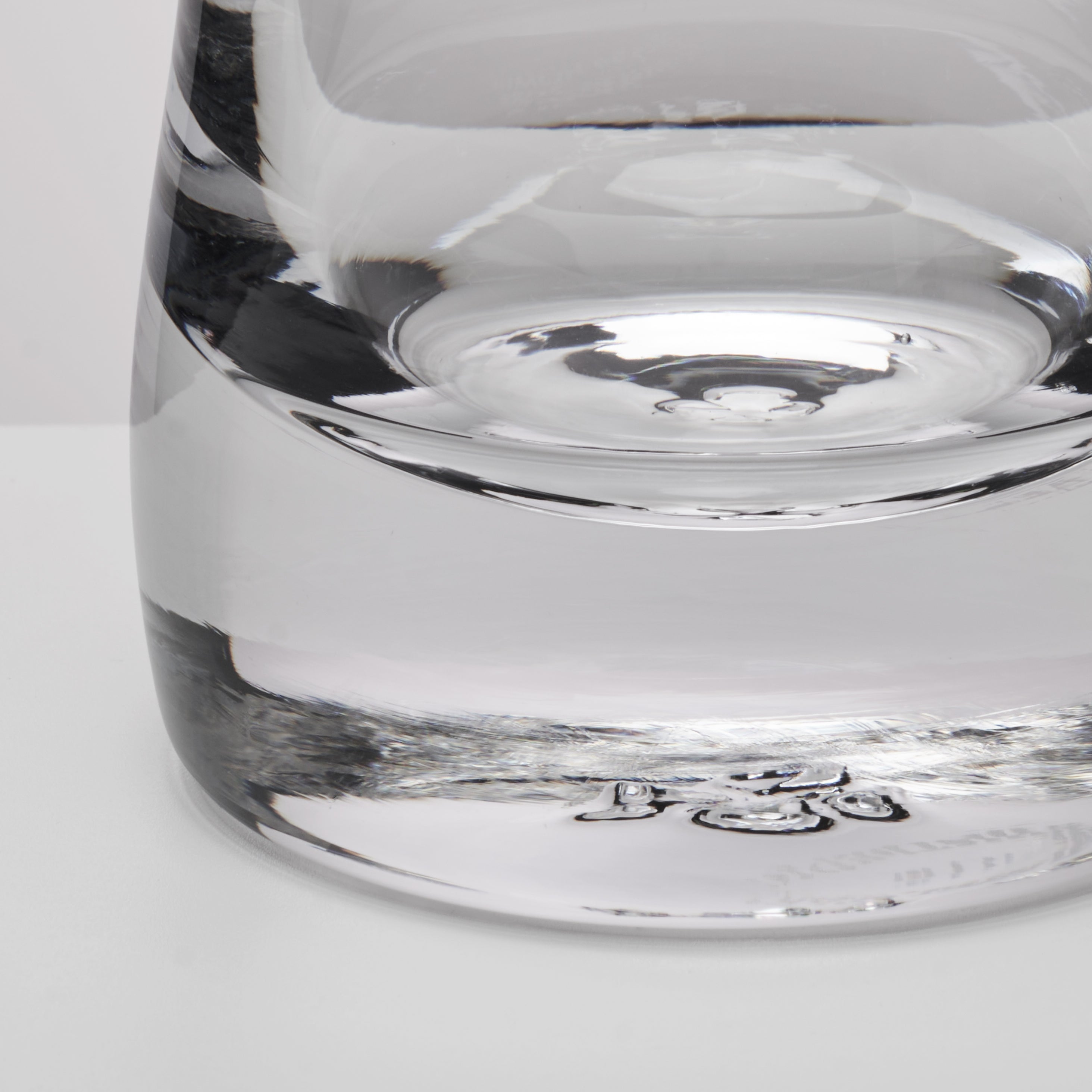 Close-up of a clear glass showing the base and lower body with reflective glass surfaces on a light gray background.