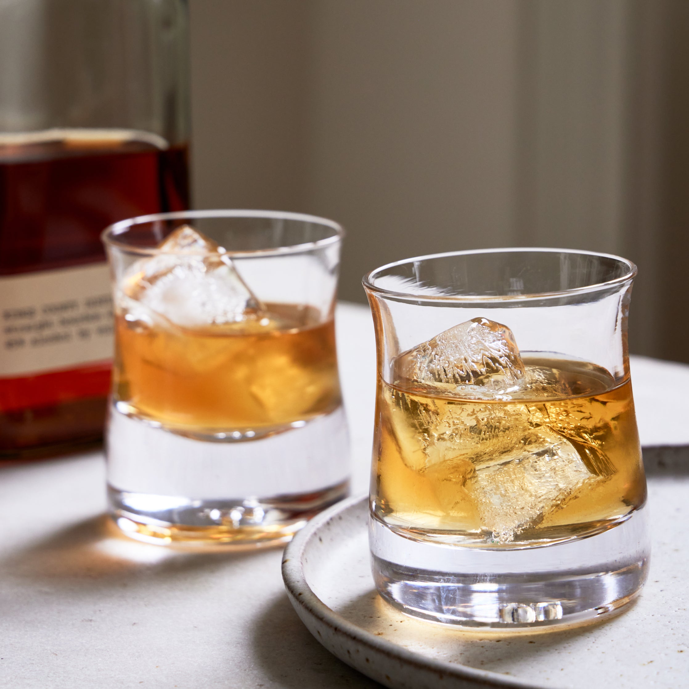Two whiskey glasses filled with amber liquid and ice cubes on a white coaster, with a bottle in the background.