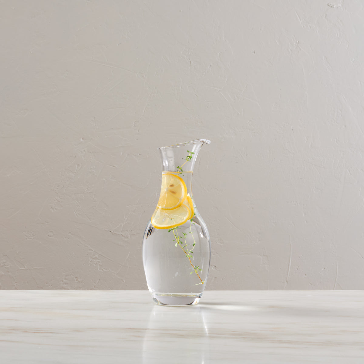 Clear glass carafe filled with water, lemon slices, and fresh herbs on a white surface.