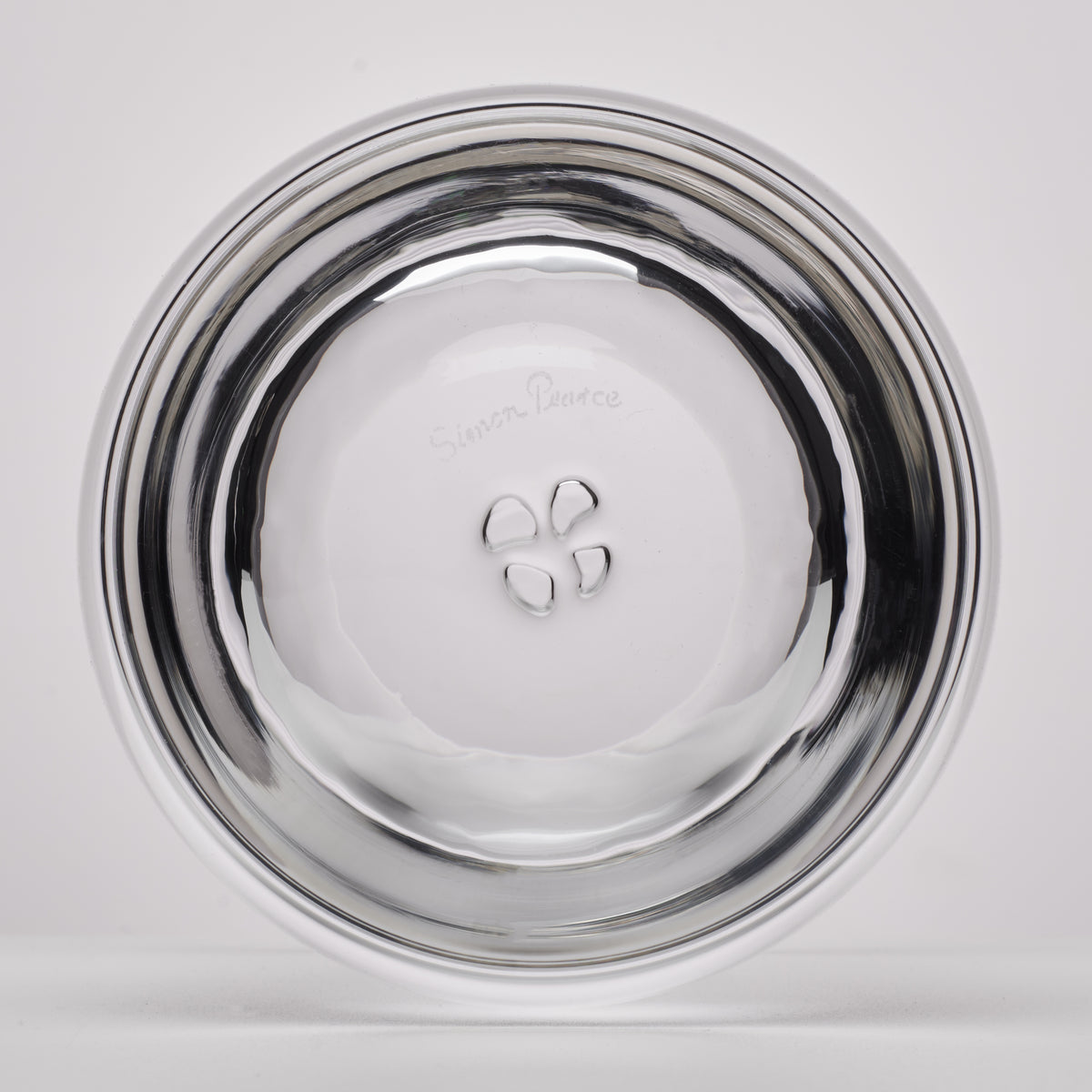 A clear glass, photographed from the bottom on a light gray background. The outside of the glass features a small embossed logo and text that appears to read 'Simon Pearce'.