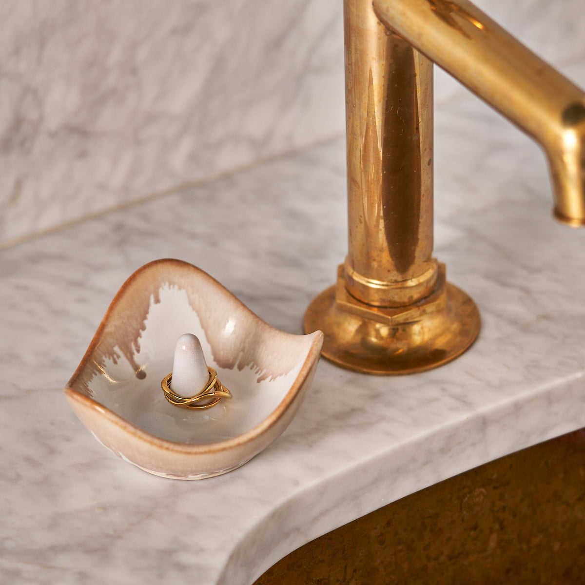 Ceramic ring holder with gold trim holding a gold ring, displayed next to a brass stand on marble surface.