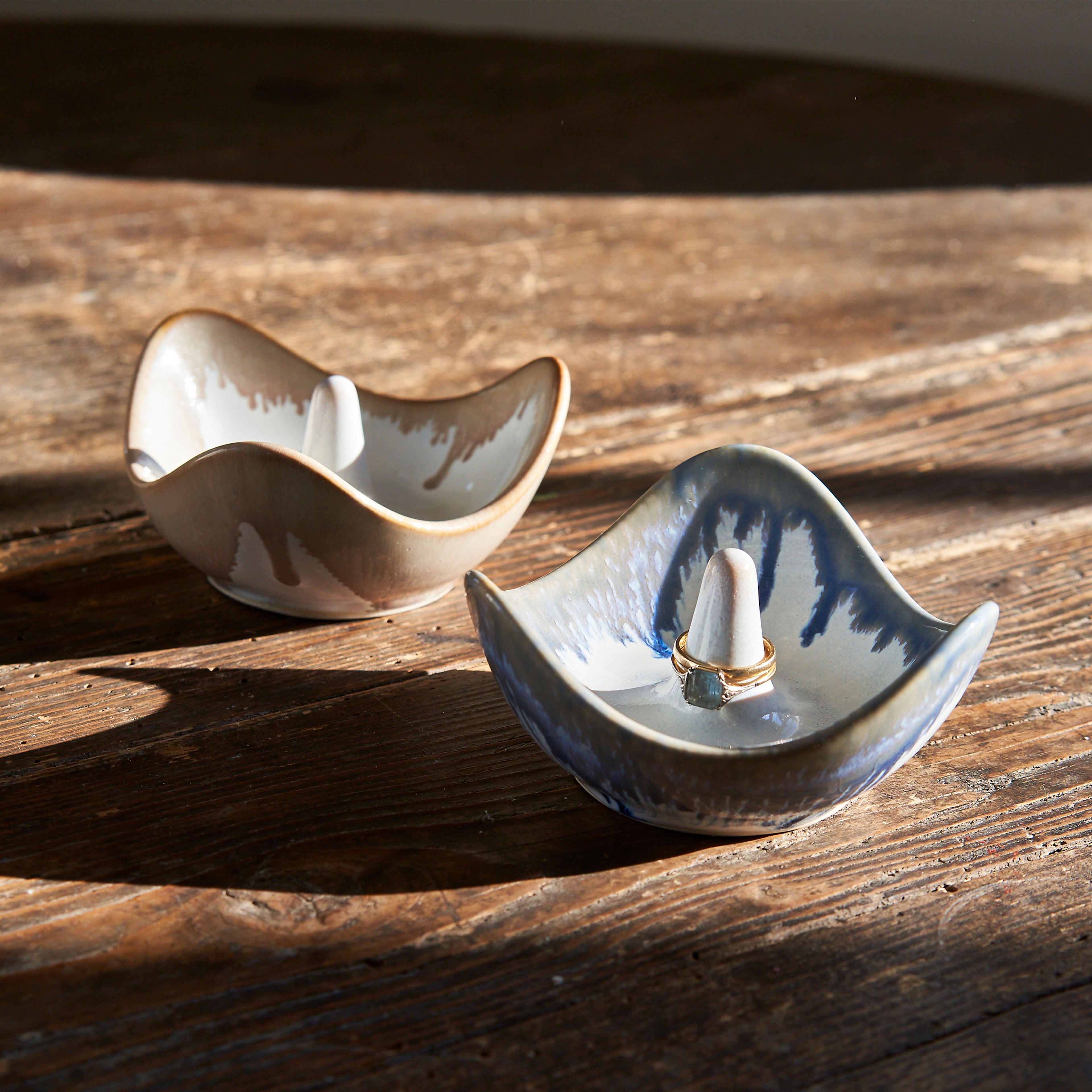 Two ceramic ring holders with curved wave shapes, one cream-colored and one blue-and-white, displayed on wooden surface.